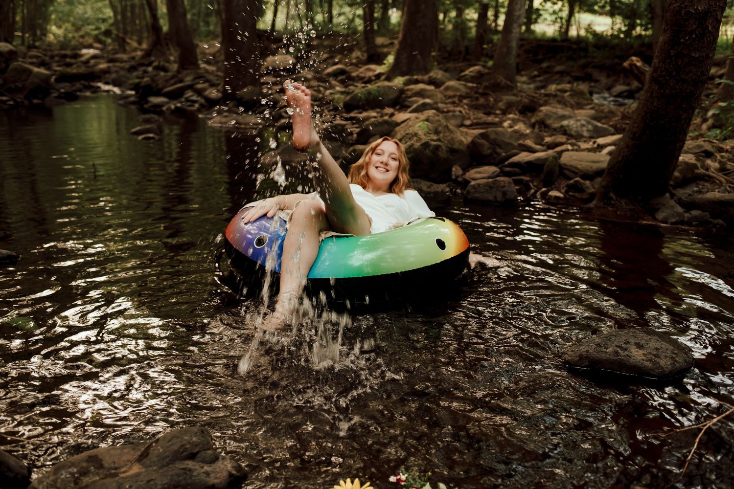 Happy young woman splashing water while floating on a colorful inflatable tube in a forest stream.