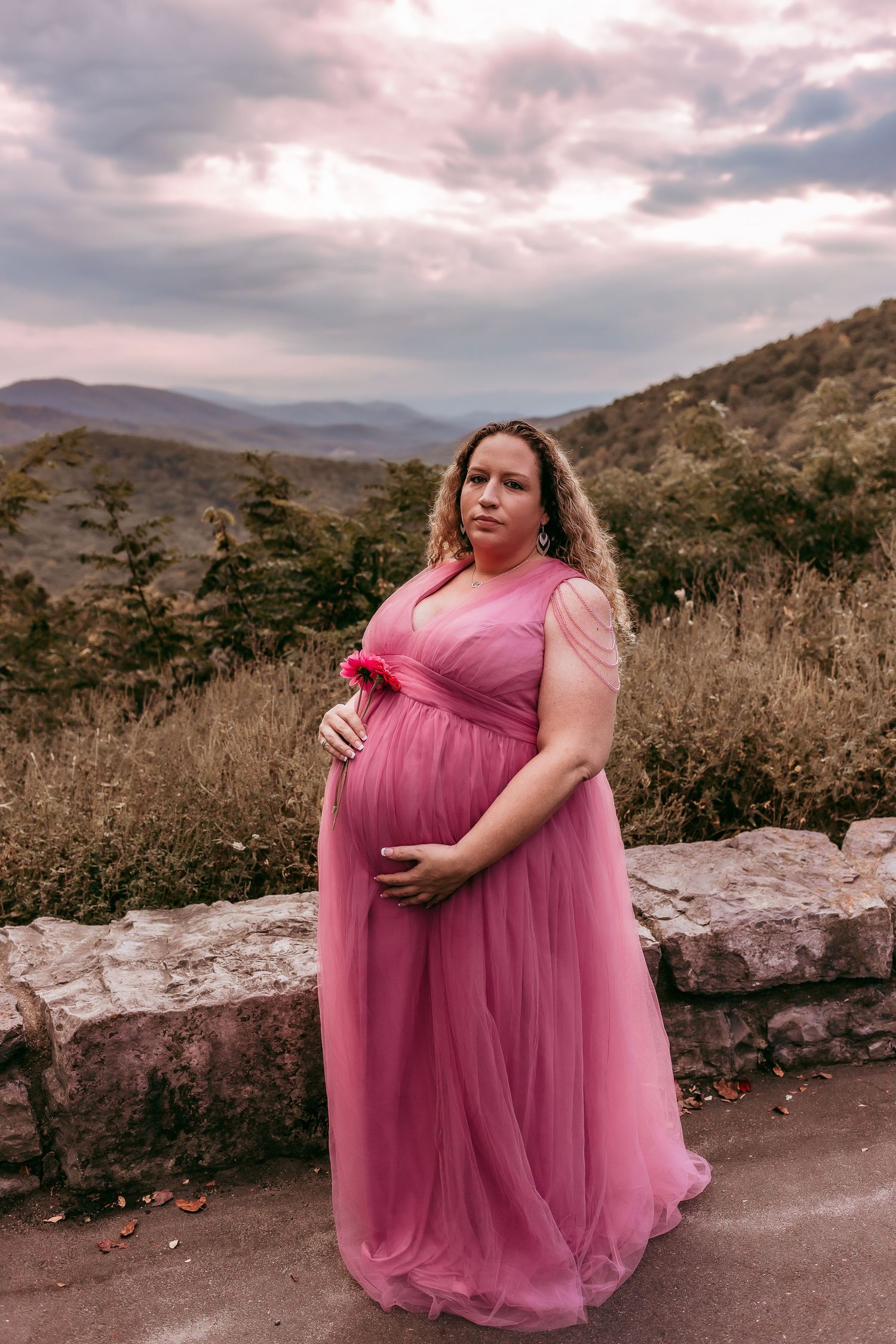 Pregnant woman in pink tulle gown cradling belly at scenic mountain overlook at dusk.