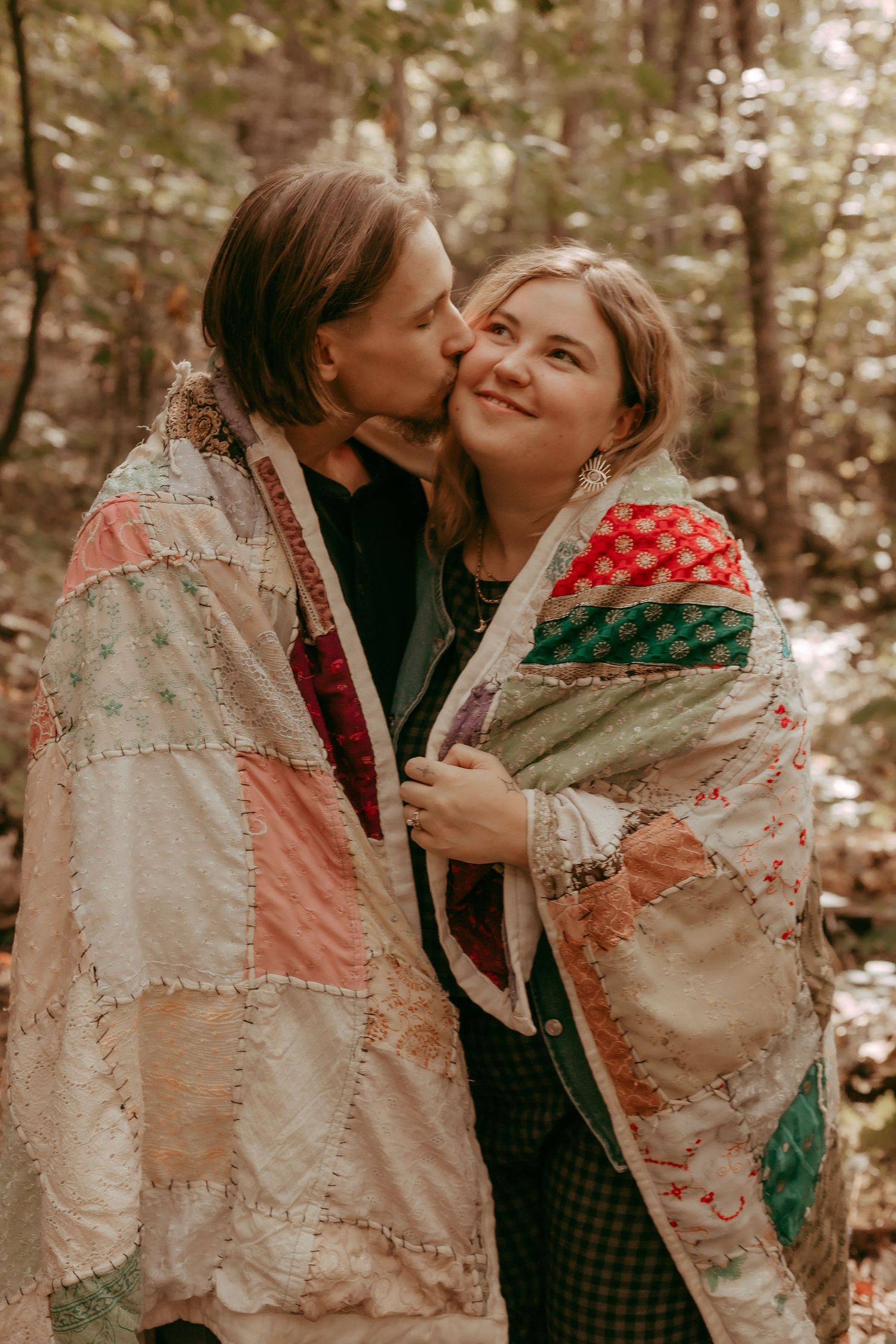 Couple wrapped in colorful patchwork quilt sharing a kiss in autumn forest.