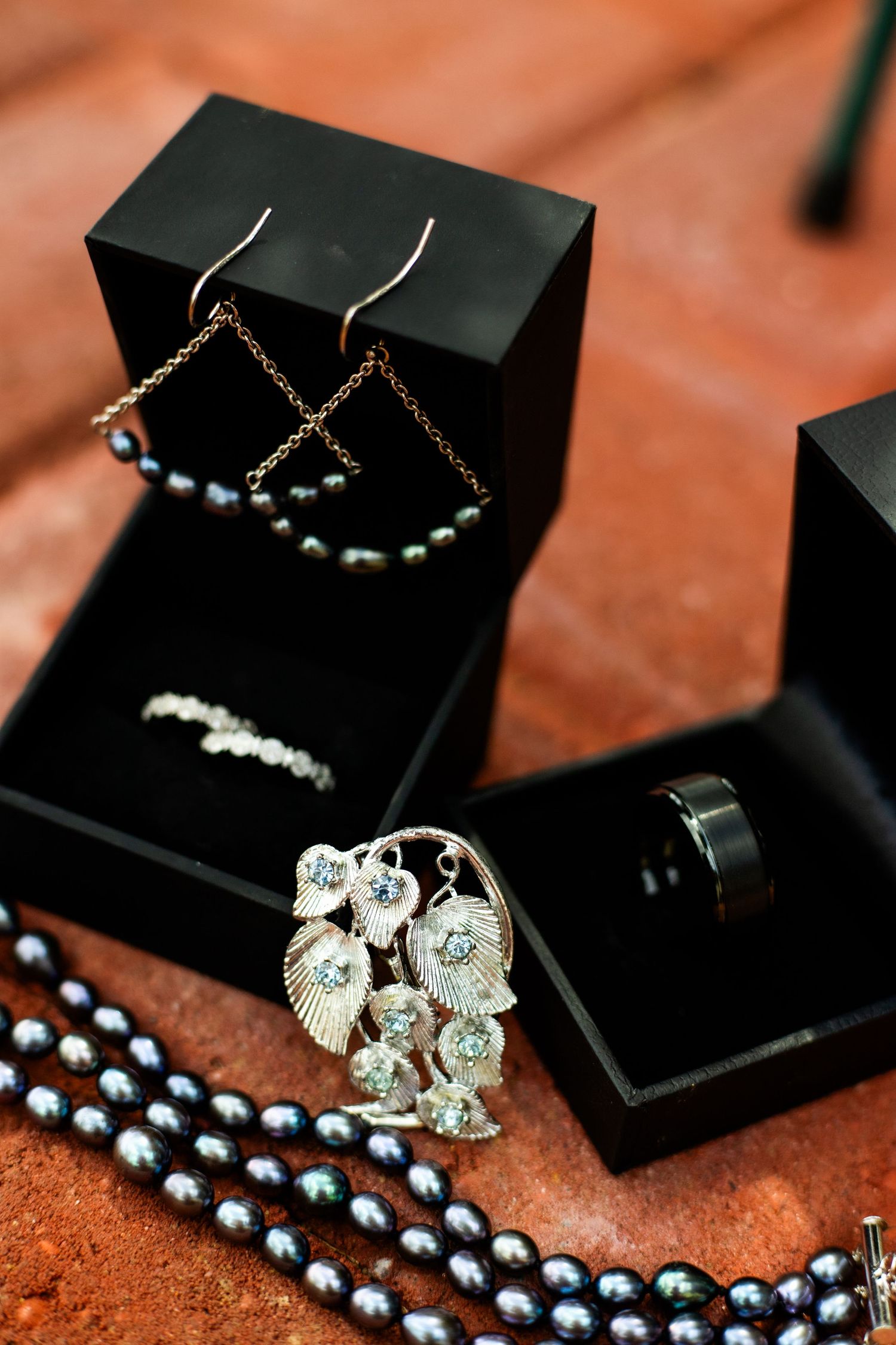 Elegant wedding jewelry display featuring pearl earrings, diamond brooch, black ring, and pearl necklace in black gift boxes.