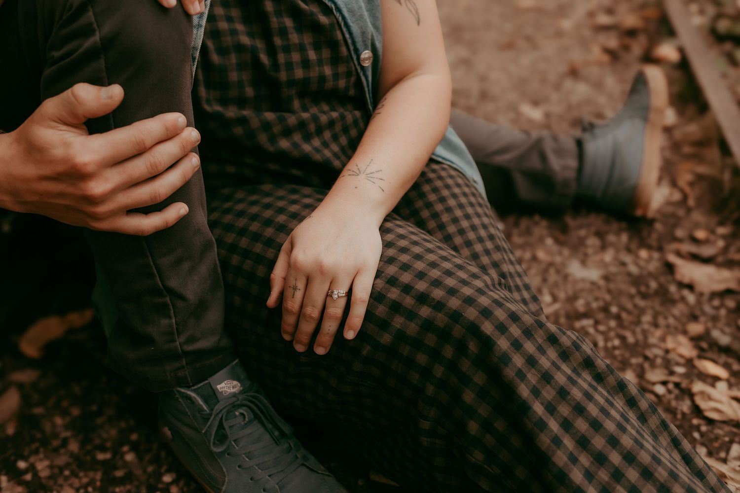 Close-up of couple holding hands showing engagement ring while sitting on forest ground.