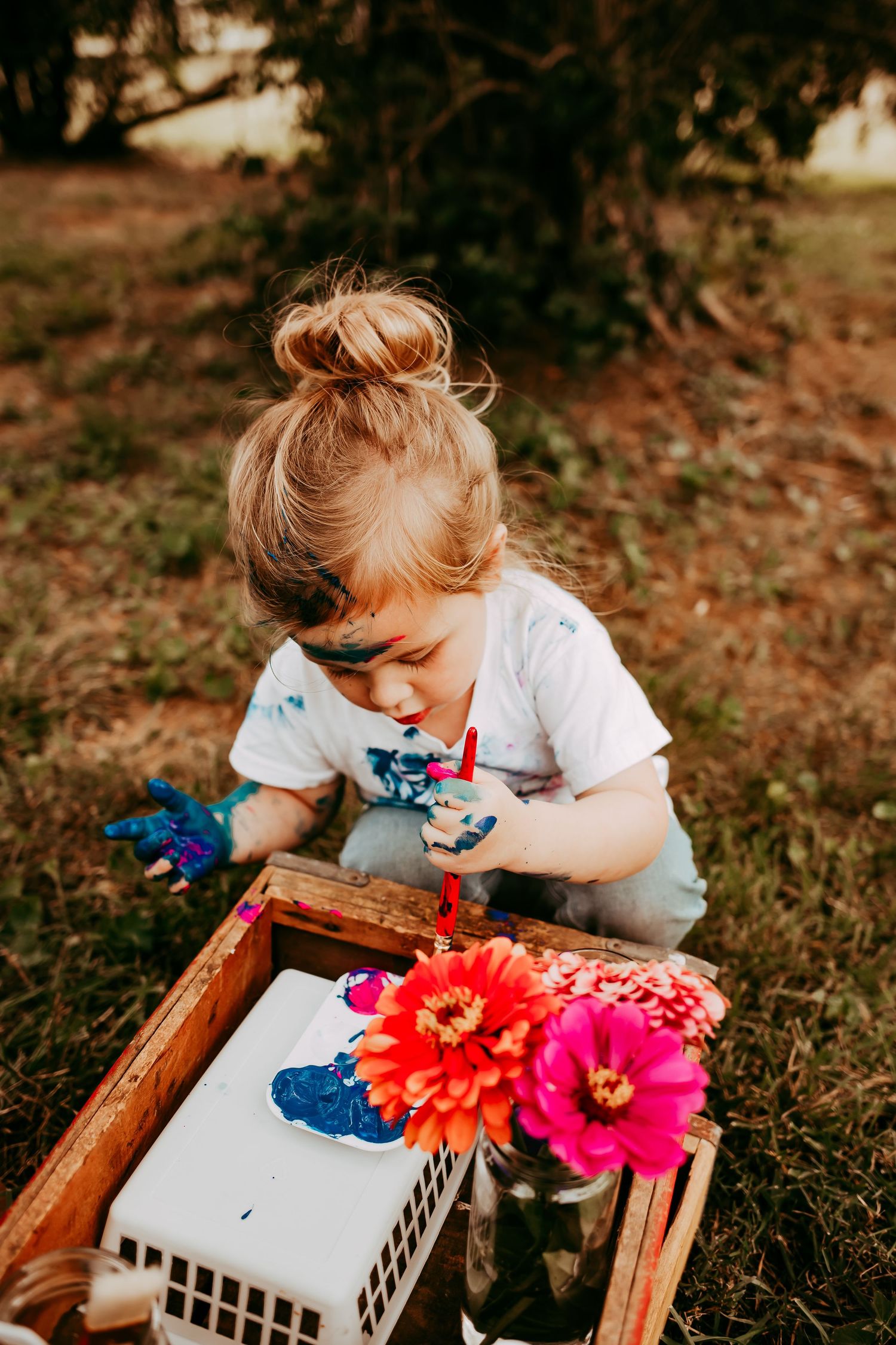 A paint-covered toddler with a bun hairstyle focuses on painting at a rustic wooden crate outdoors.