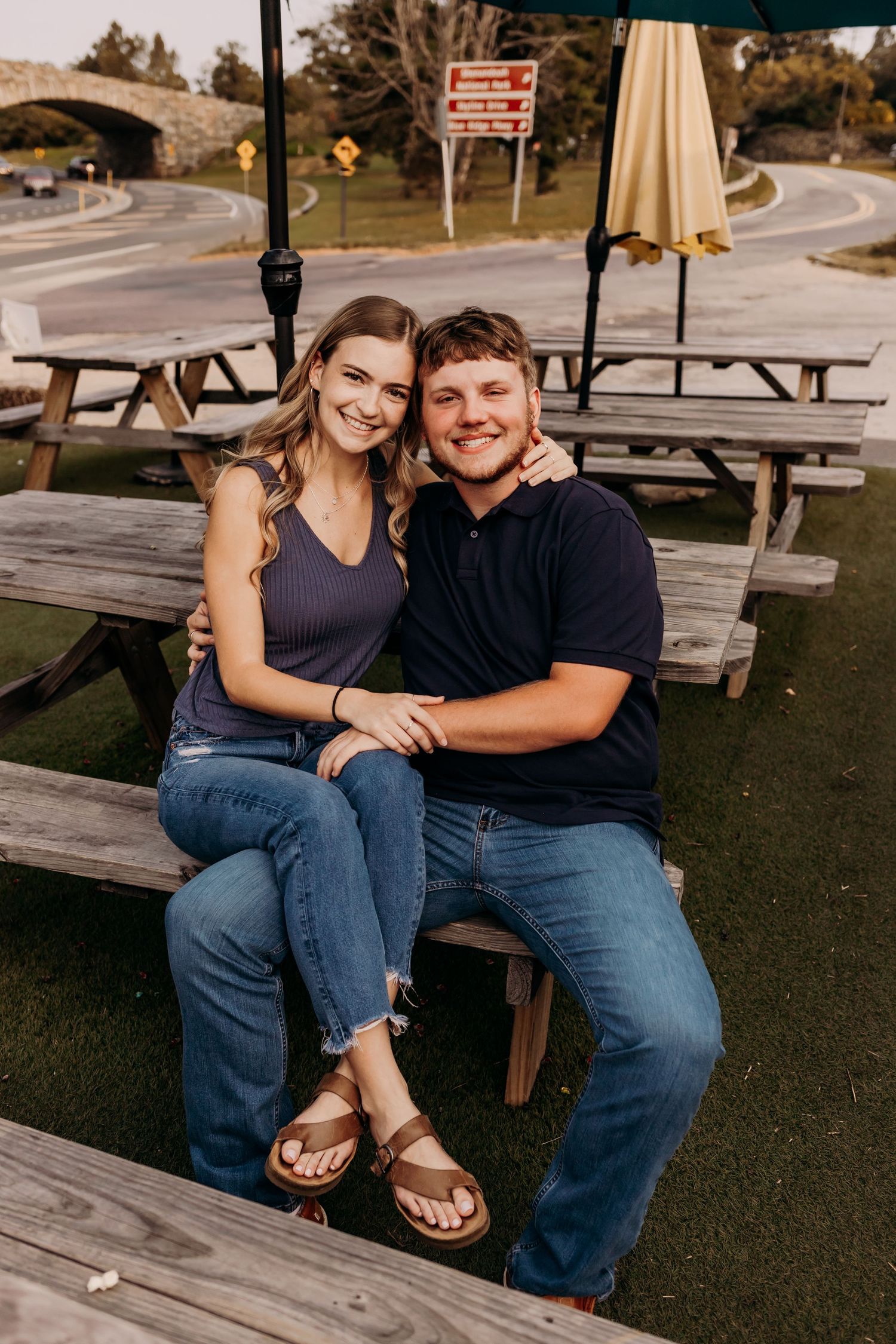 Smiling couple sitting together on a picnic bench outdoors, wearing casual jeans and tops.