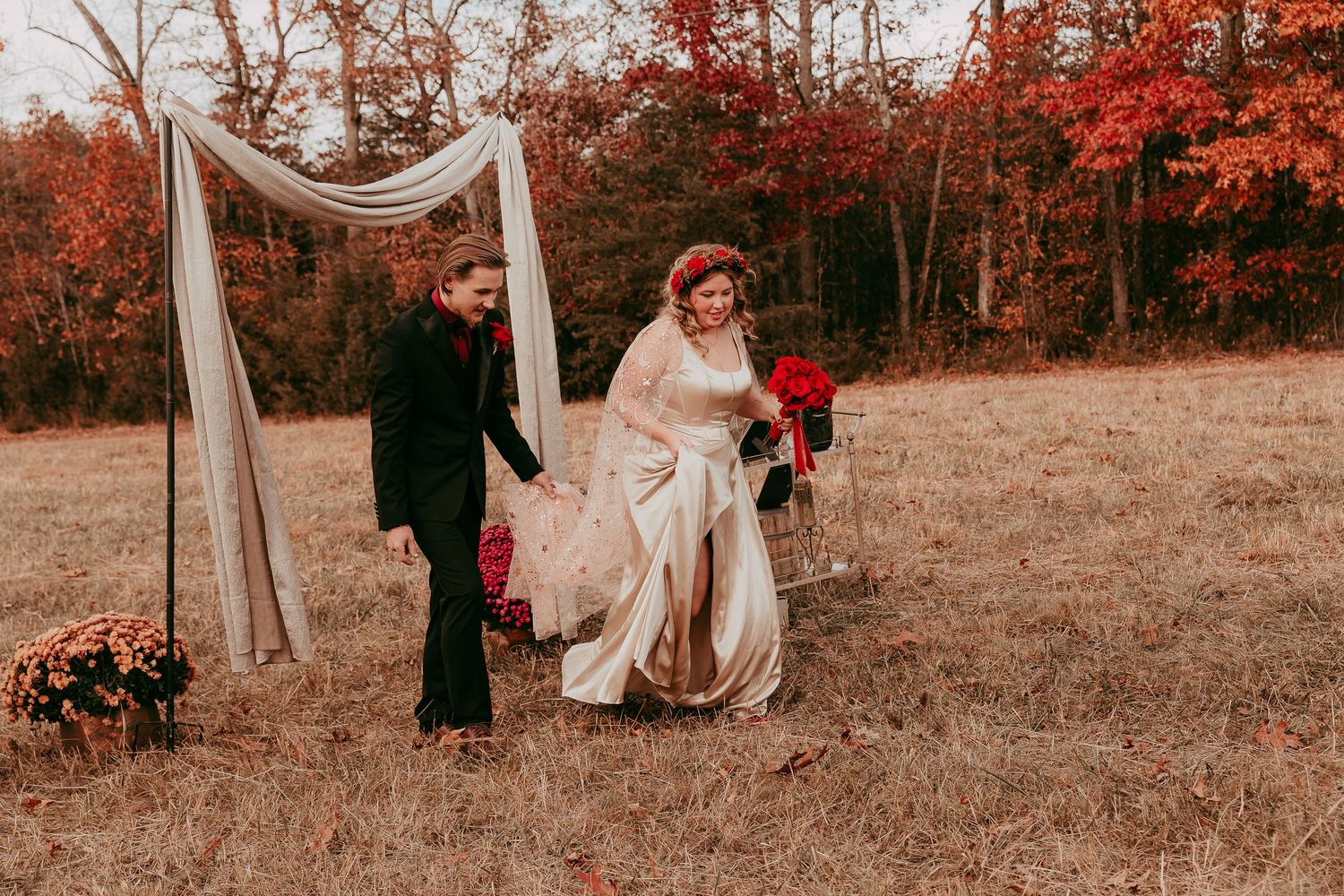 Bride in champagne gown with red bouquet and groom in black suit walking through autumn foliage at outdoor wedding.