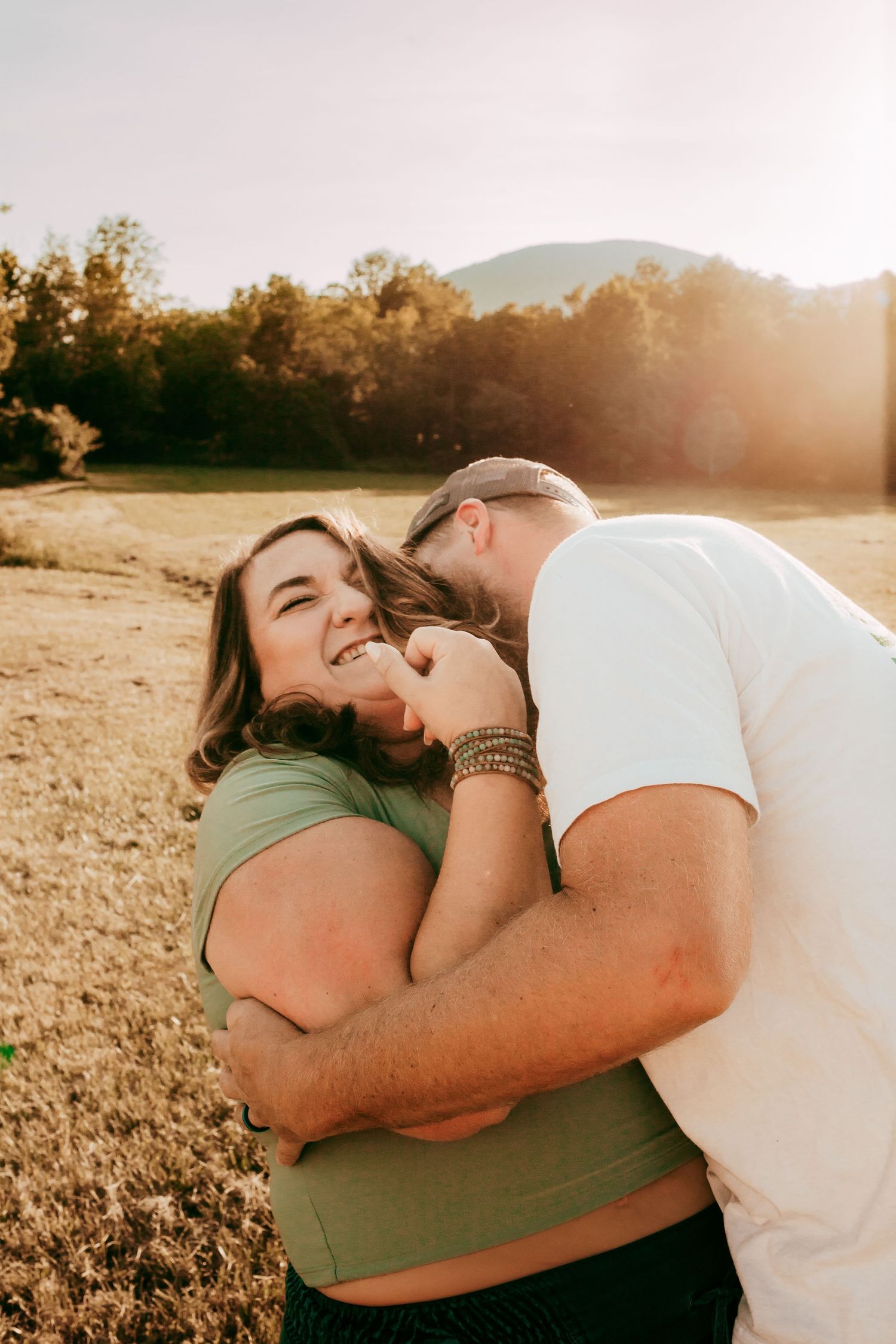 Couple sharing playful intimate moment in golden sunlight in open meadow with mountain backdrop.
