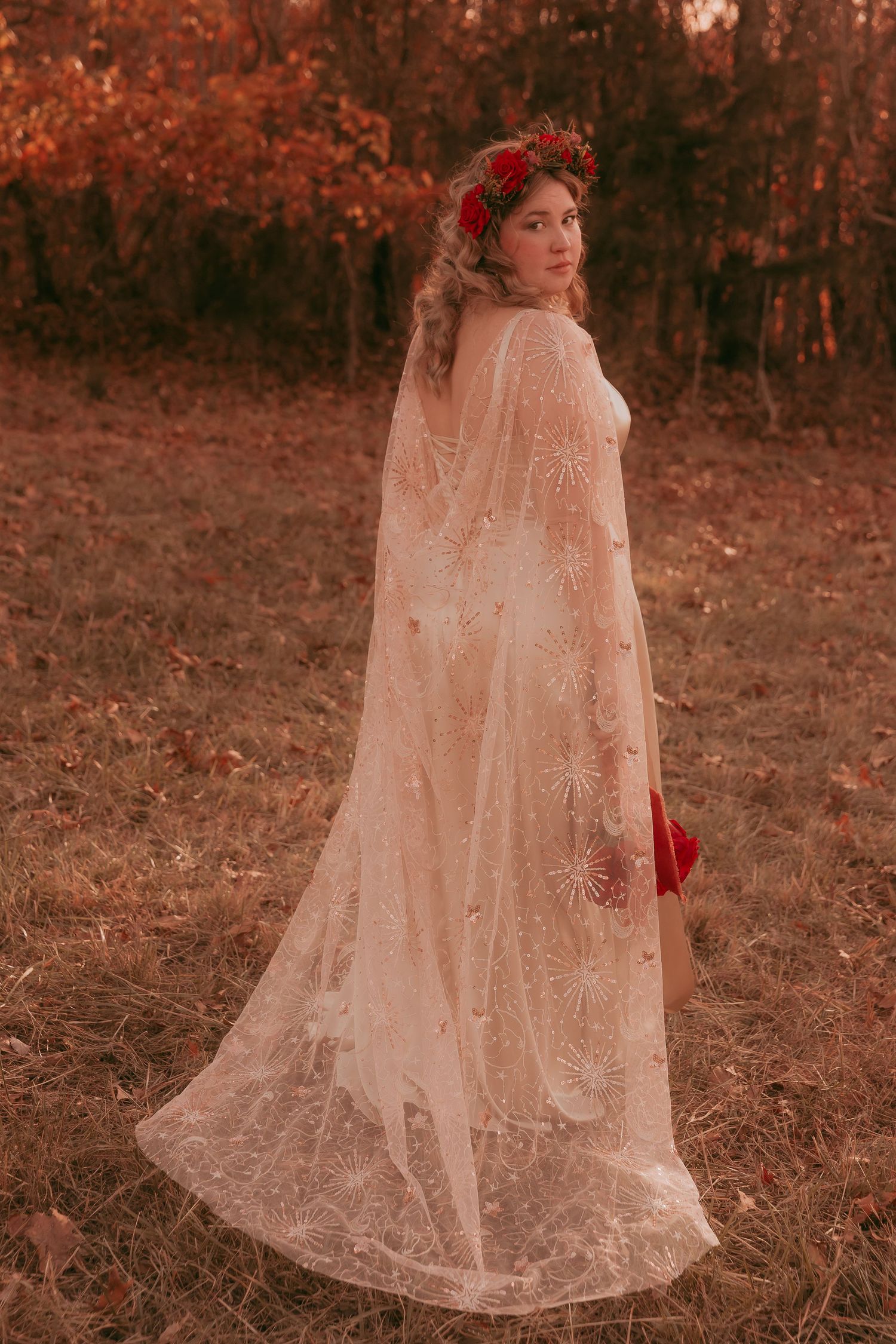 Bride in flowing gold lace cape and red floral crown poses in autumn woodland.