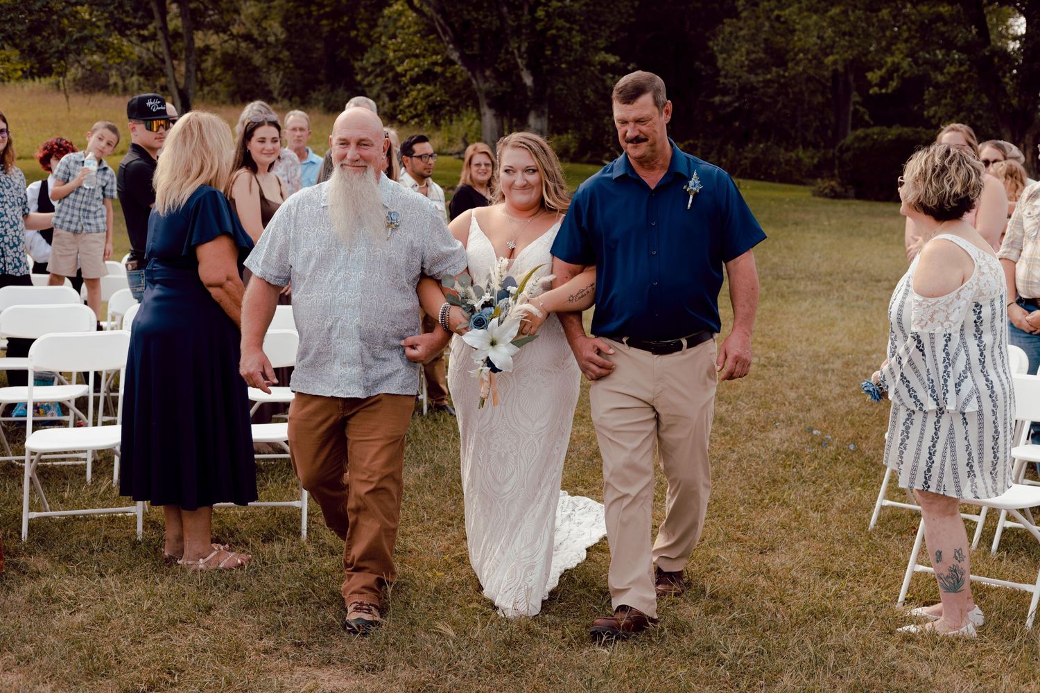 Bride in lace wedding gown being walked down the aisle by two men at an outdoor wedding.
