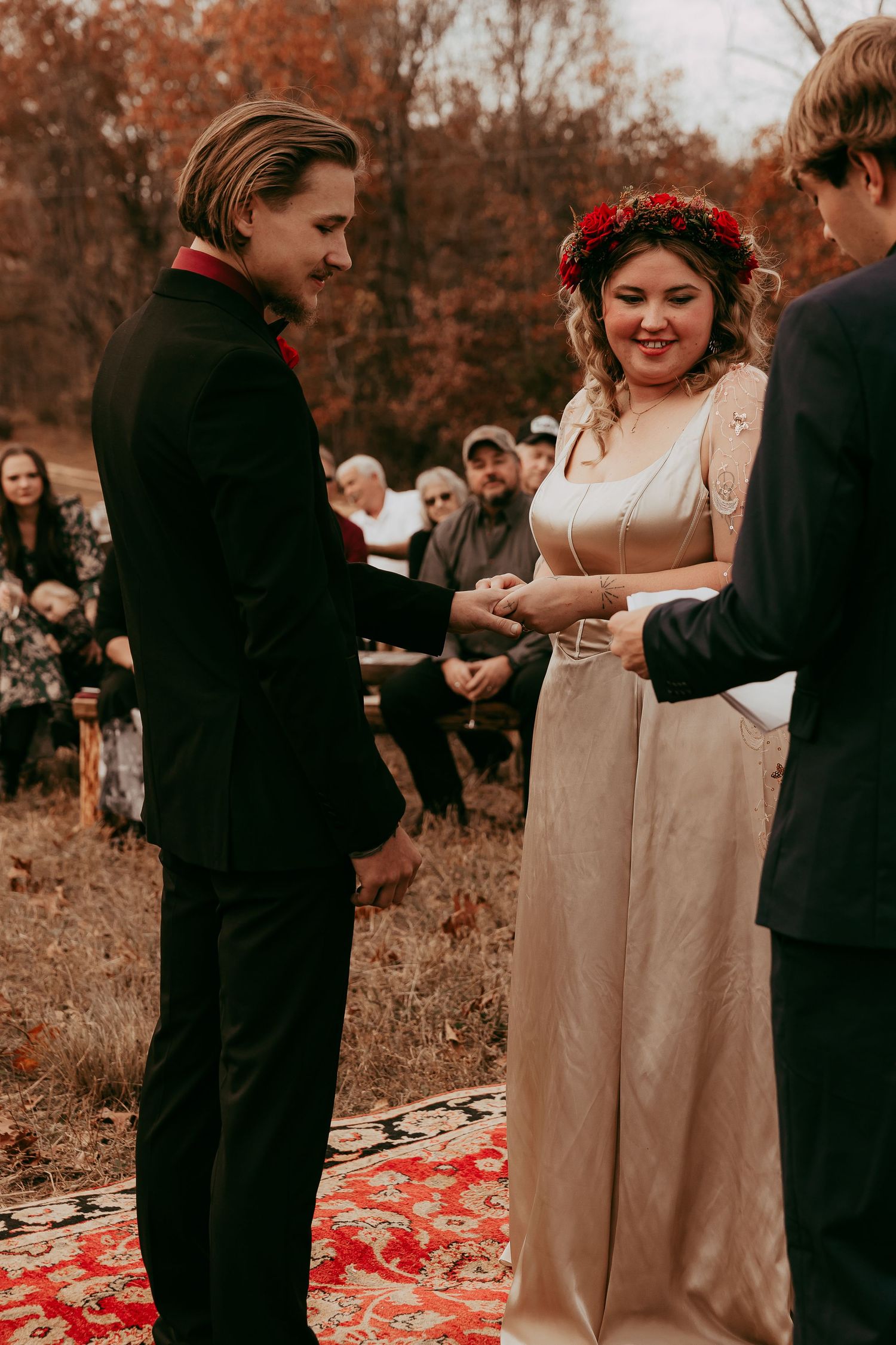 Couple exchanges rings during outdoor fall wedding ceremony on red patterned rug.