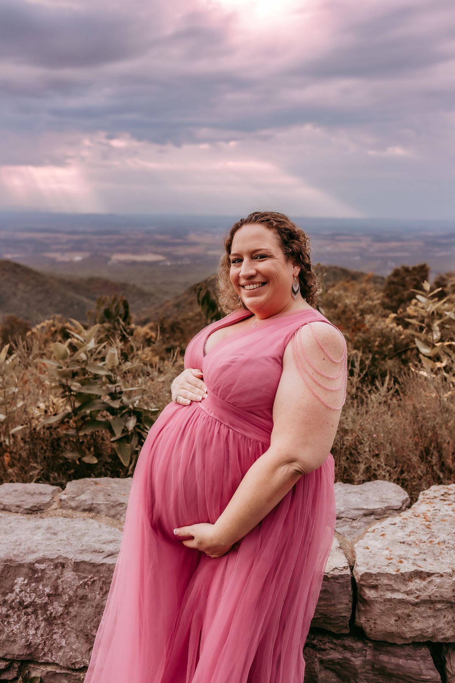 Smiling pregnant woman in pink tulle gown at mountain overlook with dramatic cloudy sky.