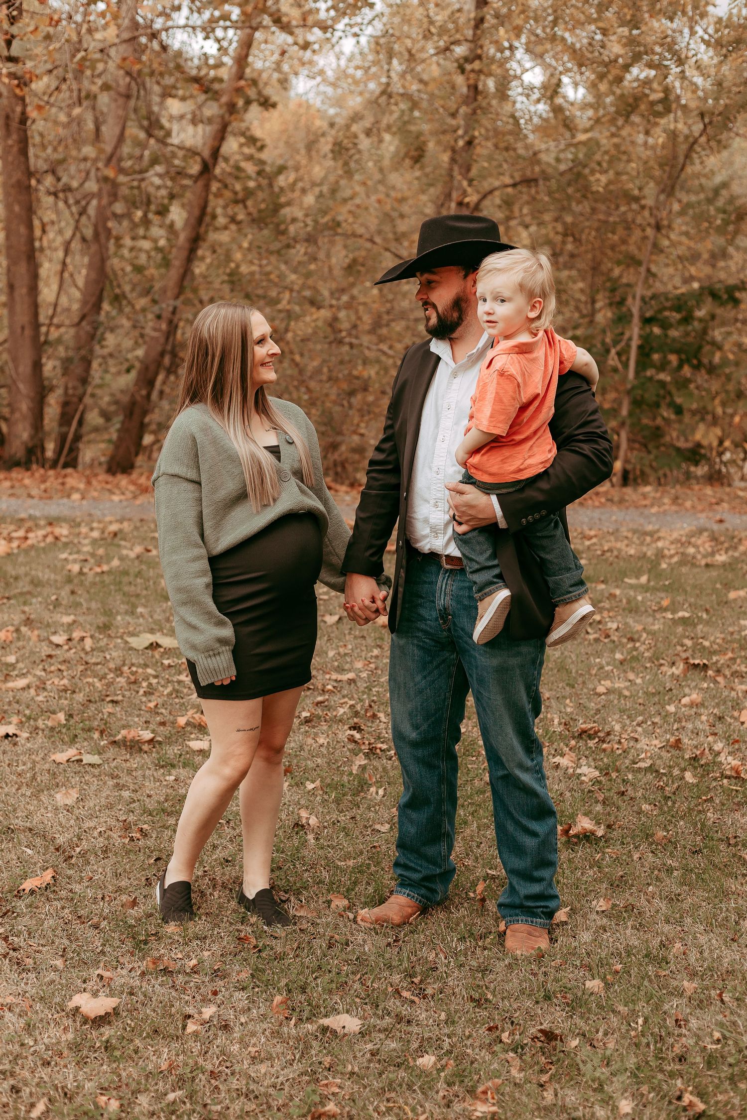 Pregnant woman and cowboy hat man holding toddler boy hand-in-hand in fall foliage park.
