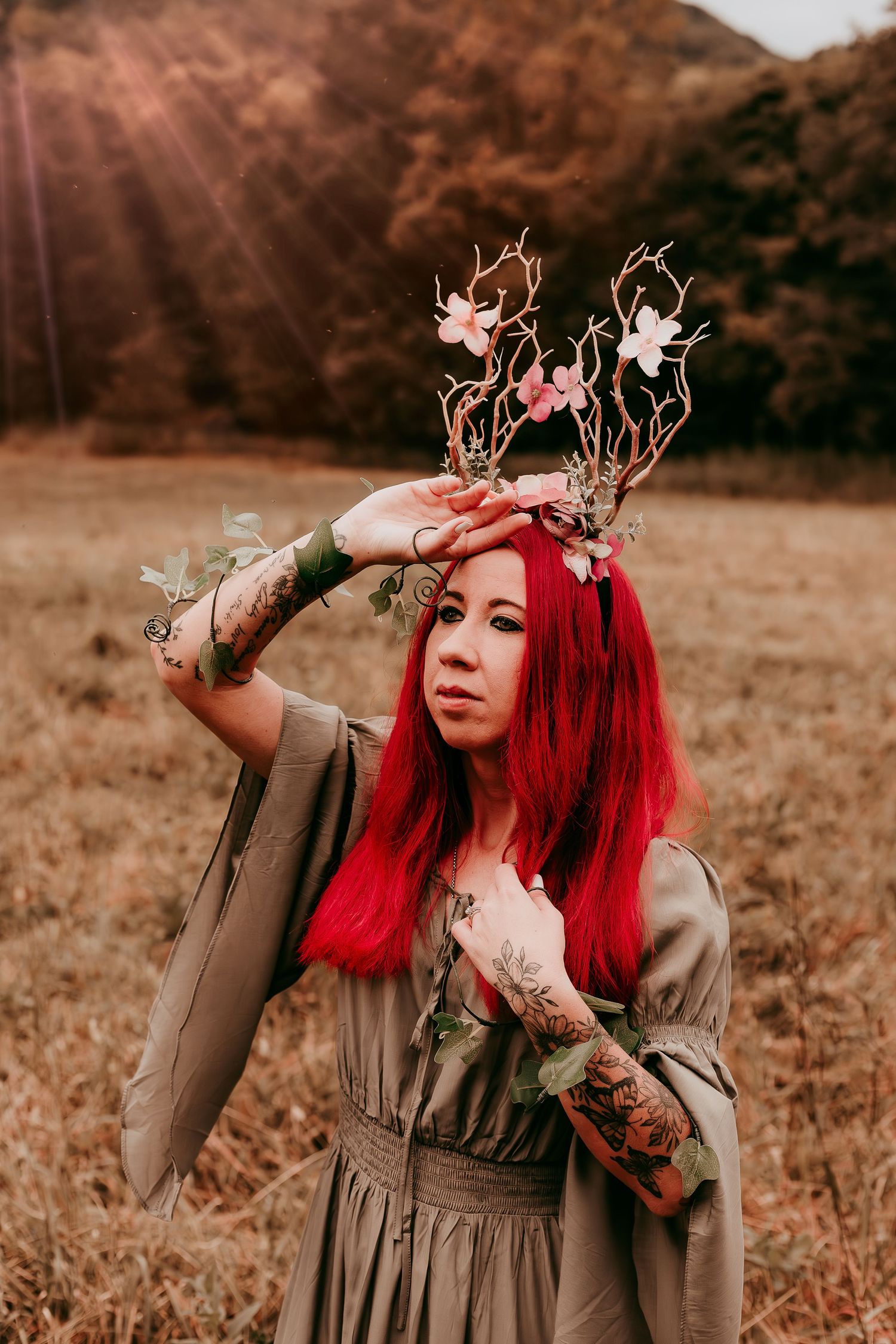 Woman with red hair and tattoos wearing floral antler crown in golden autumn field with sunrays.