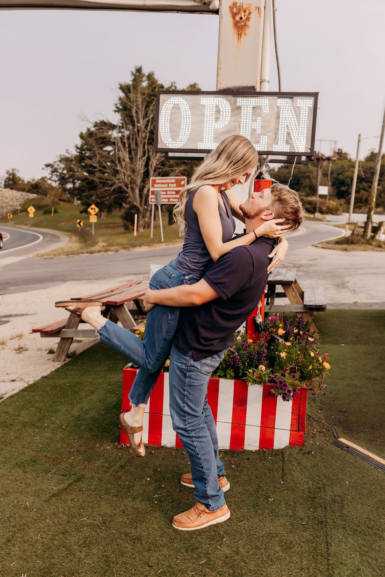 Couple sharing a romantic kiss as man lifts woman near vintage OPEN sign at roadside diner.