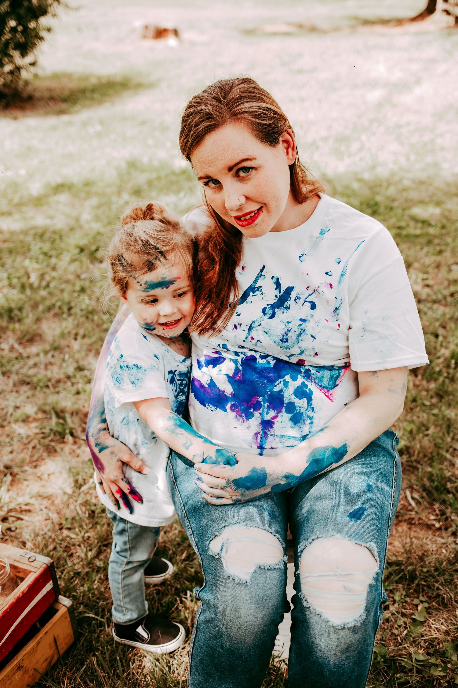 A smiling pregnant woman covered in colorful paint poses with her paint-splattered toddler in a grassy yard.