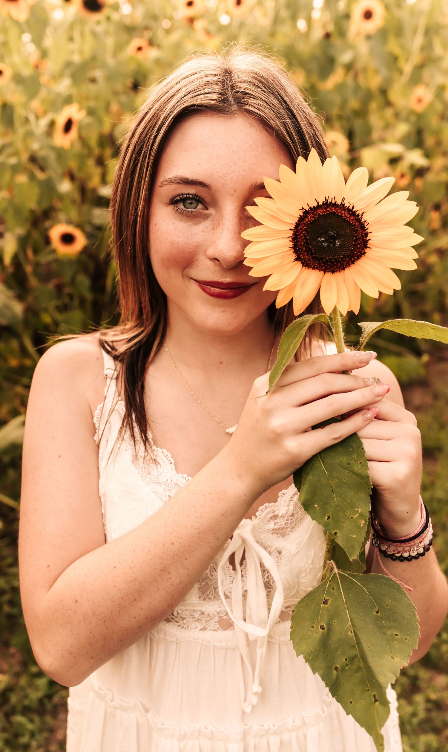 Smiling young woman with freckles holds sunflower over eye in sunflower field wearing white dress.