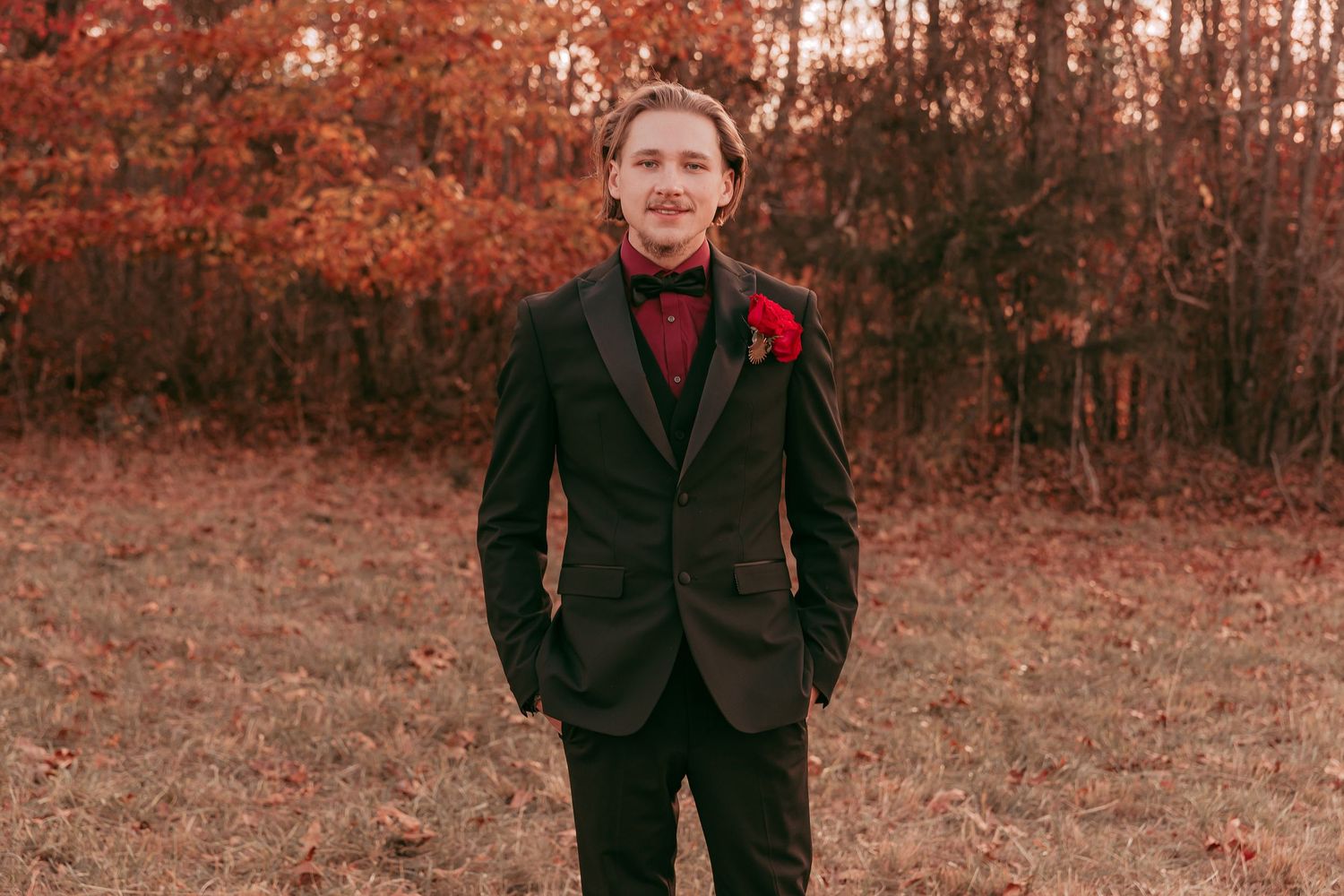 Groom in black tuxedo with red boutonniere and bow tie stands in fall foliage.