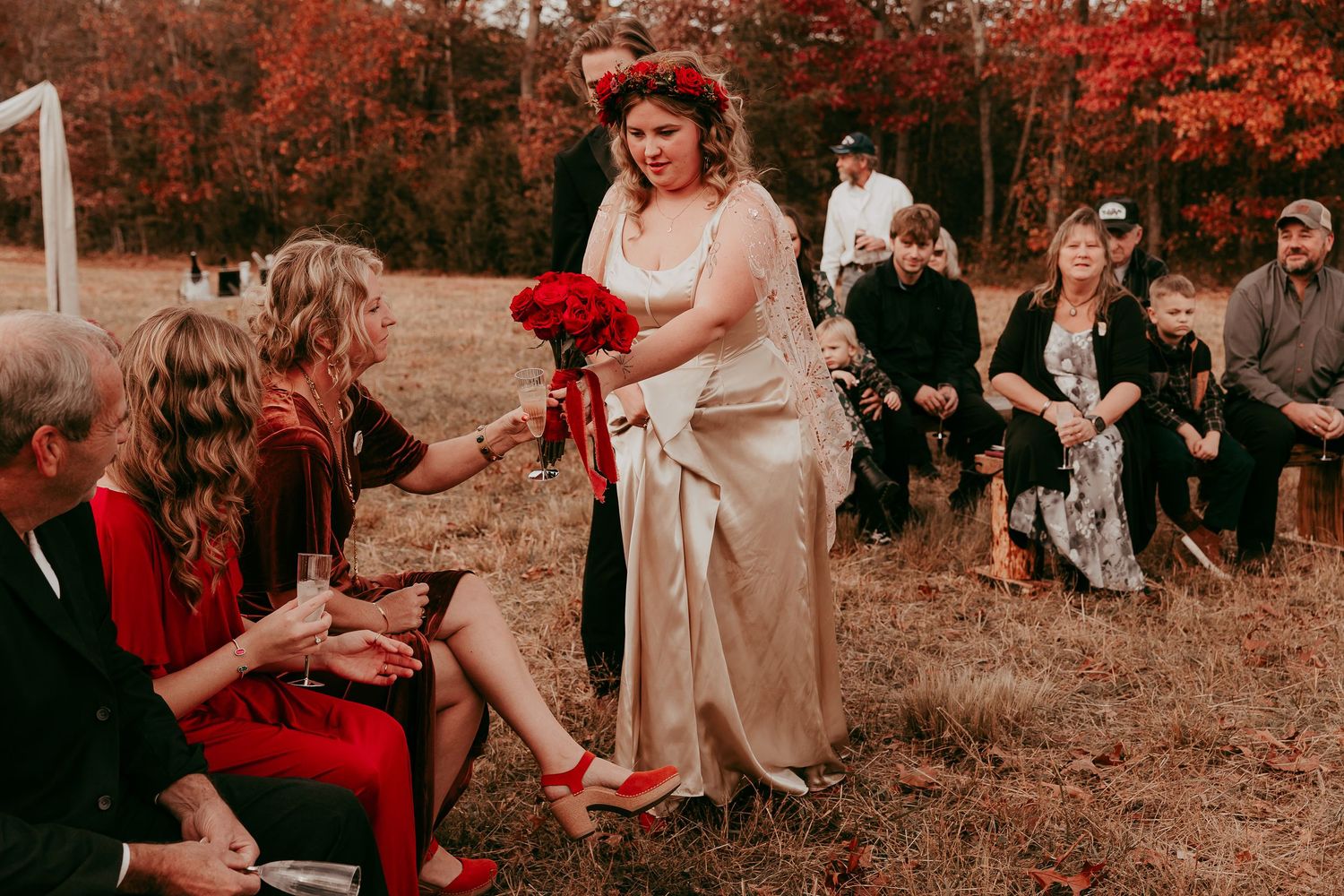 Bride in champagne satin gown with red floral crown greeting seated guests at intimate outdoor fall wedding ceremony.