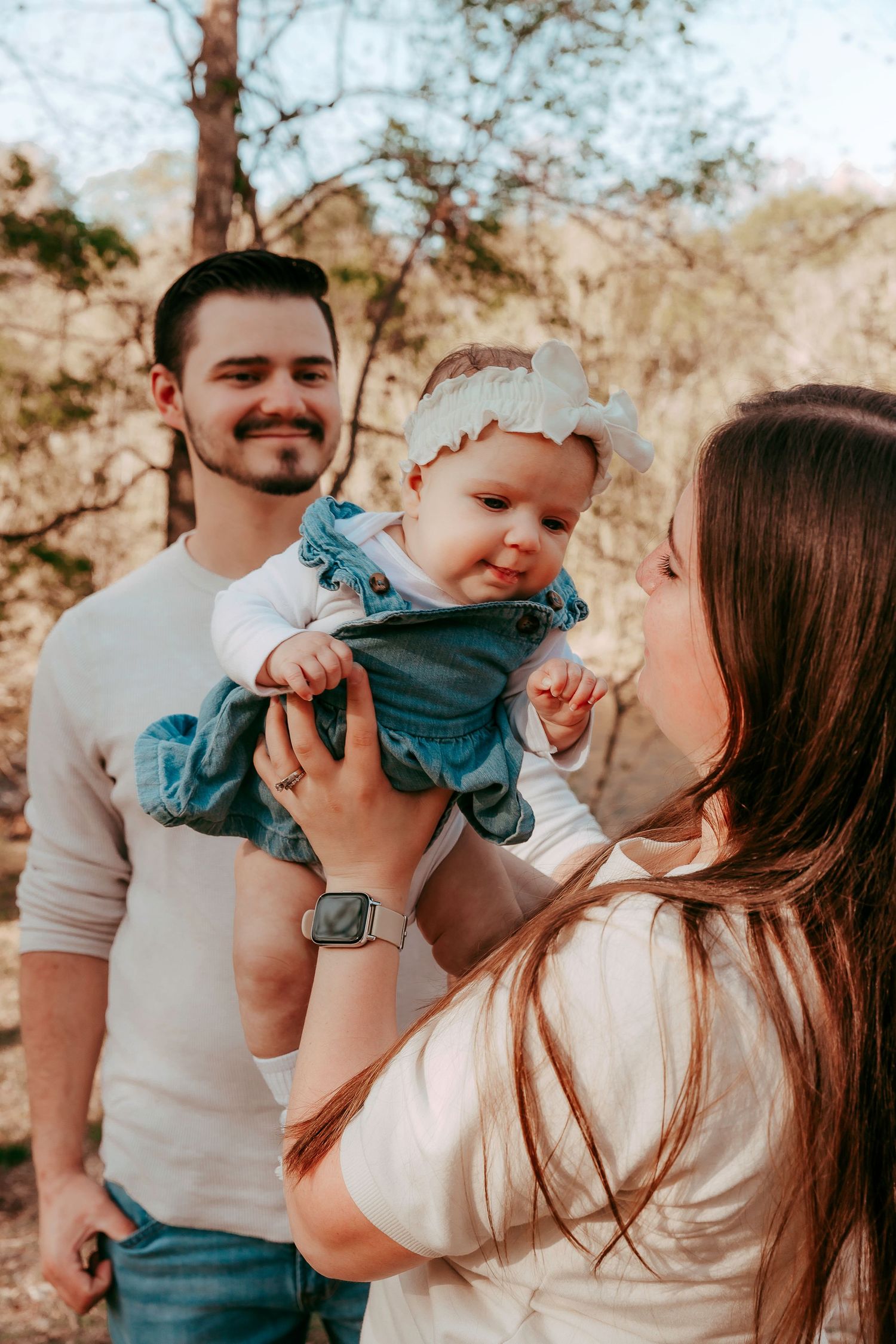 A young family outdoors, parents holding their smiling baby girl in a blue dress and white headband among trees.