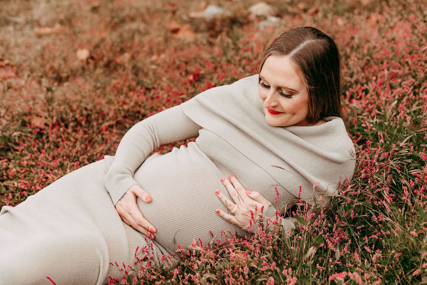 Pregnant woman in beige dress lying in field of red wildflowers, cradling baby bump with red lips.