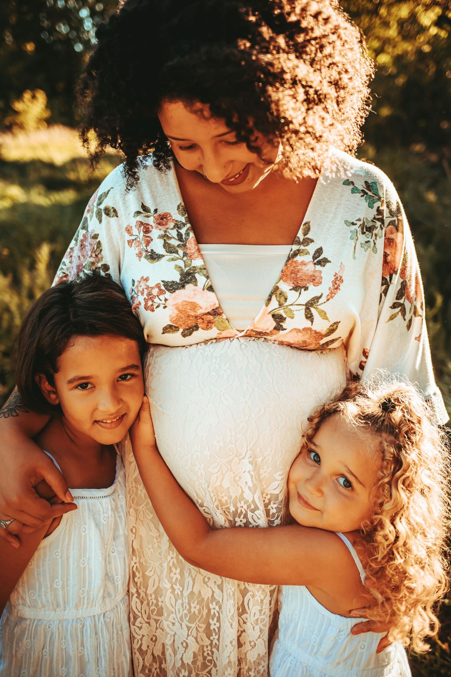 Pregnant woman with curly hair in floral kimono embraced by two smiling girls outdoors.