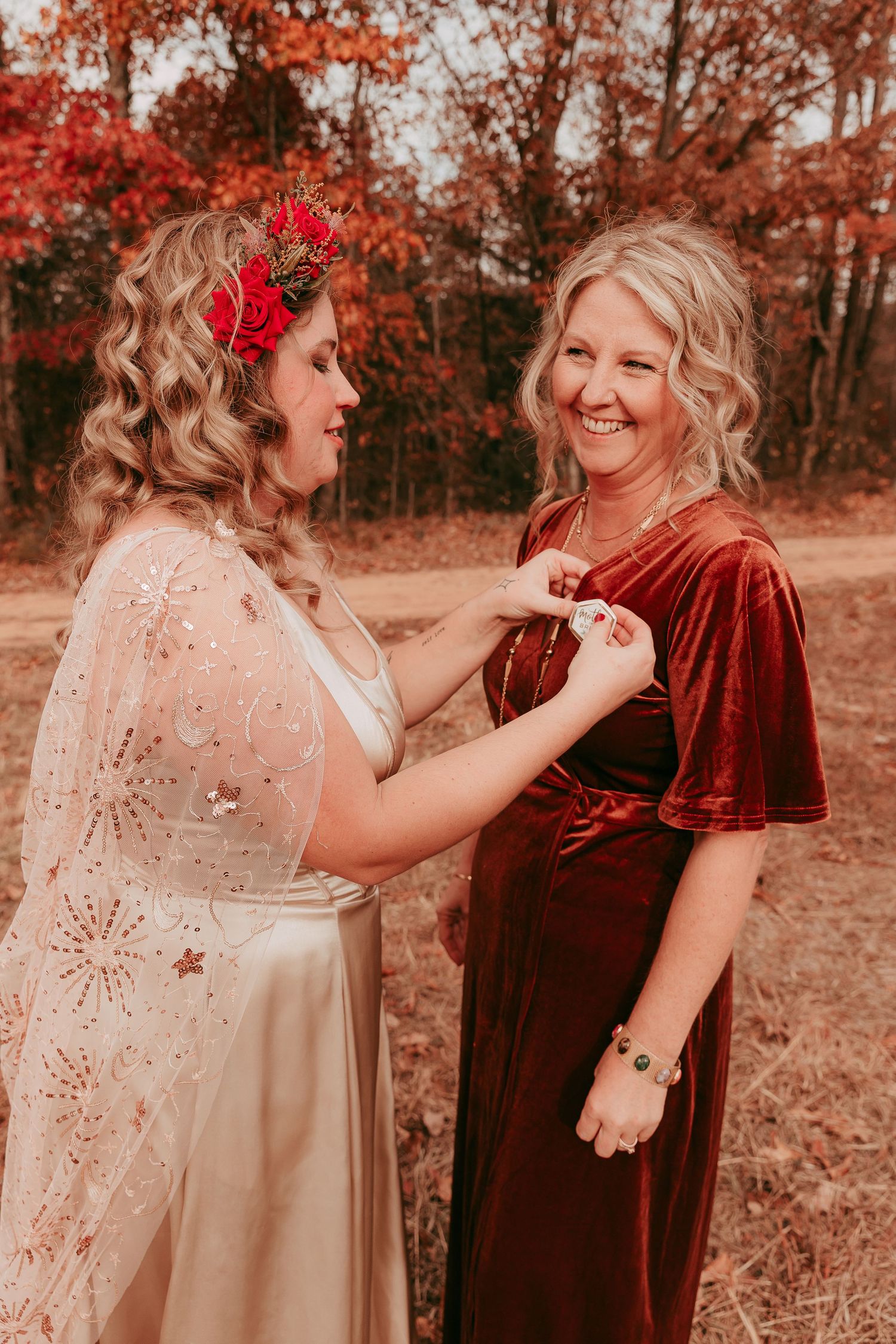 Bride in gold lace cape adjusts bridesmaid's burgundy velvet dress amid autumn foliage.