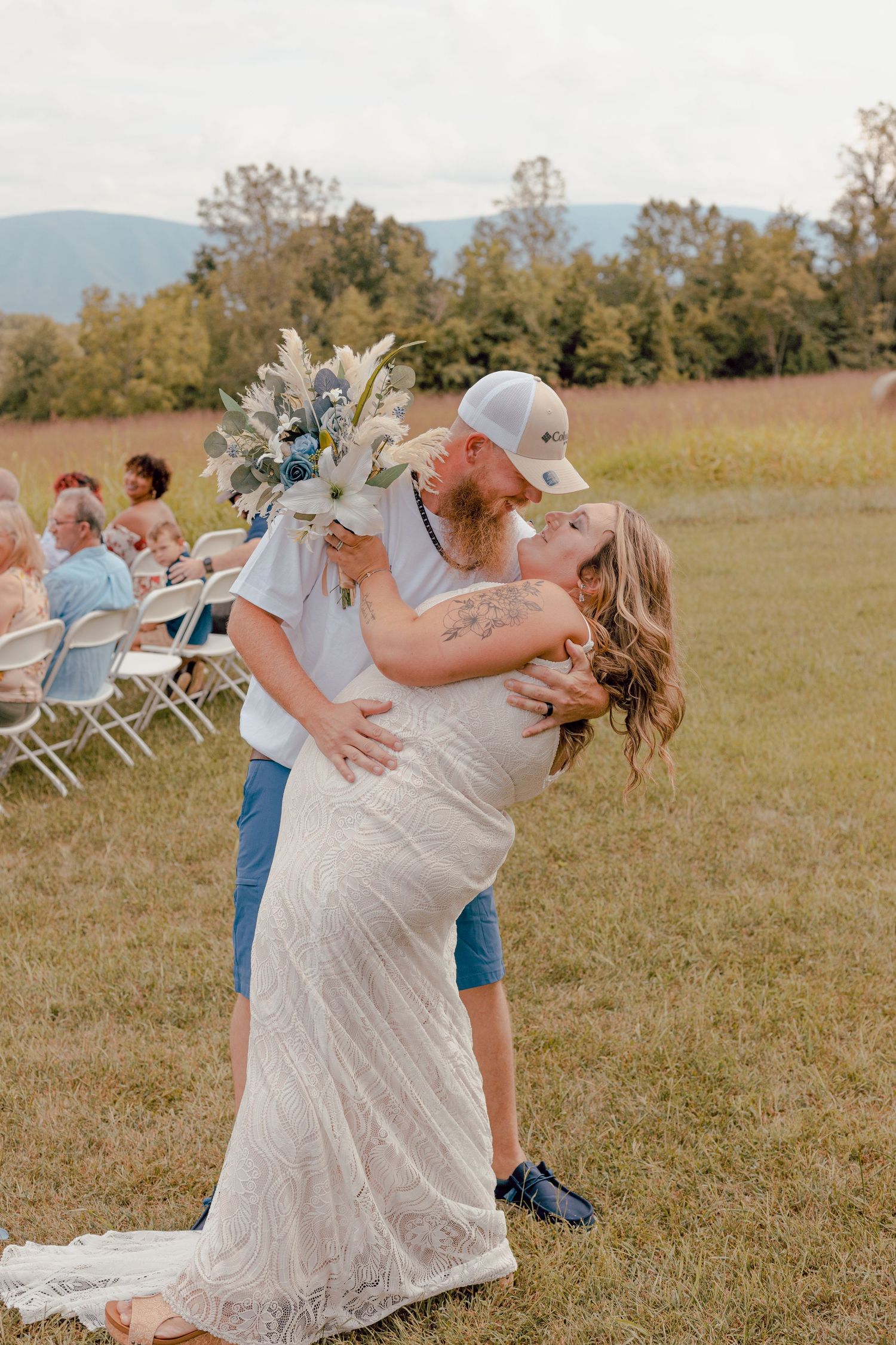 Bride and groom share a romantic kiss outdoors at a rustic field wedding, bride holding a white and blue bouquet.