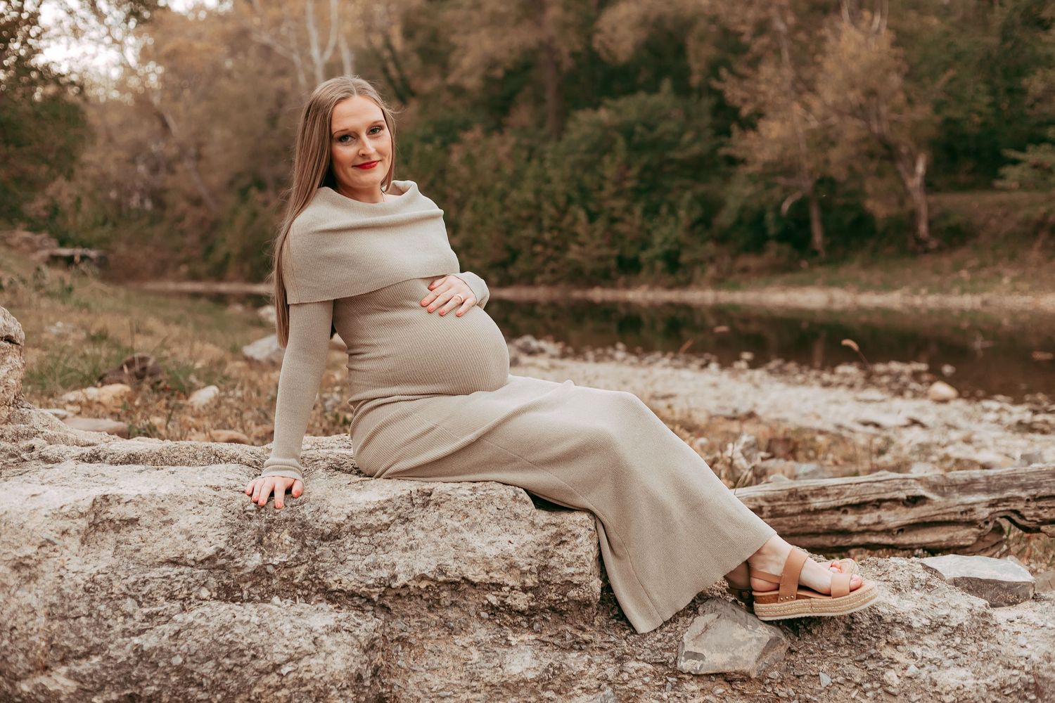 Pregnant woman in tan knit dress sits on rocks near a river in an autumn landscape.