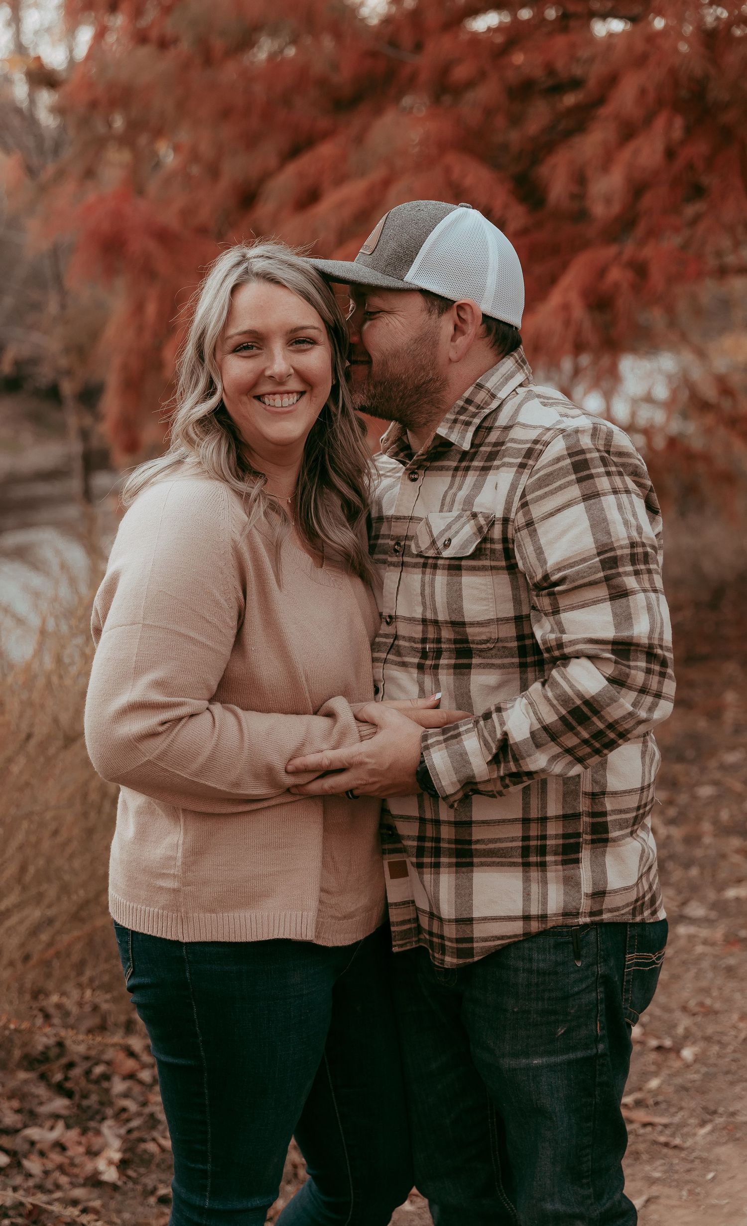 Smiling couple embraces and shares a kiss among brilliant red fall foliage outdoors.
