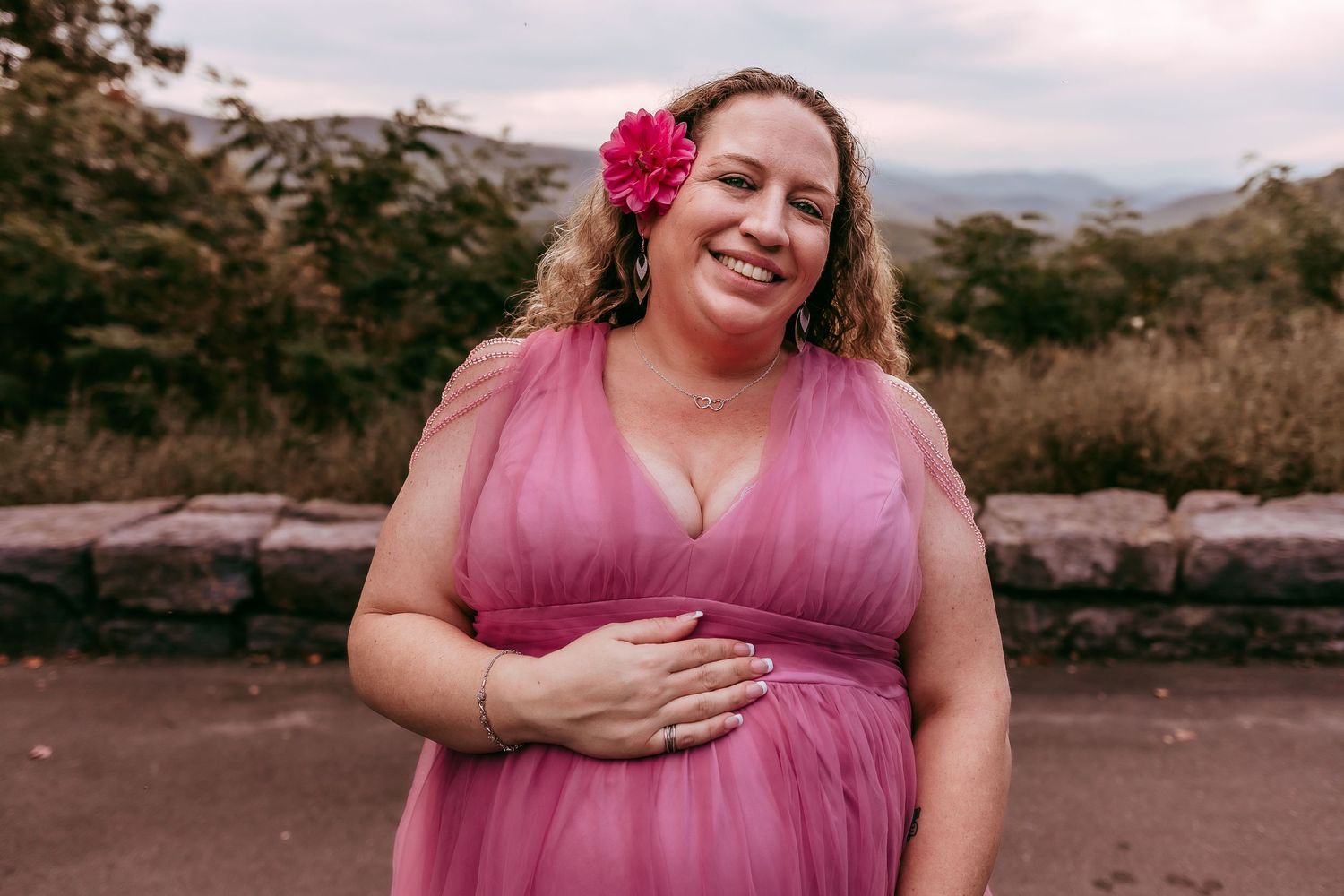 Smiling pregnant woman in pink tulle dress with flower in hair poses outdoors with mountain views.