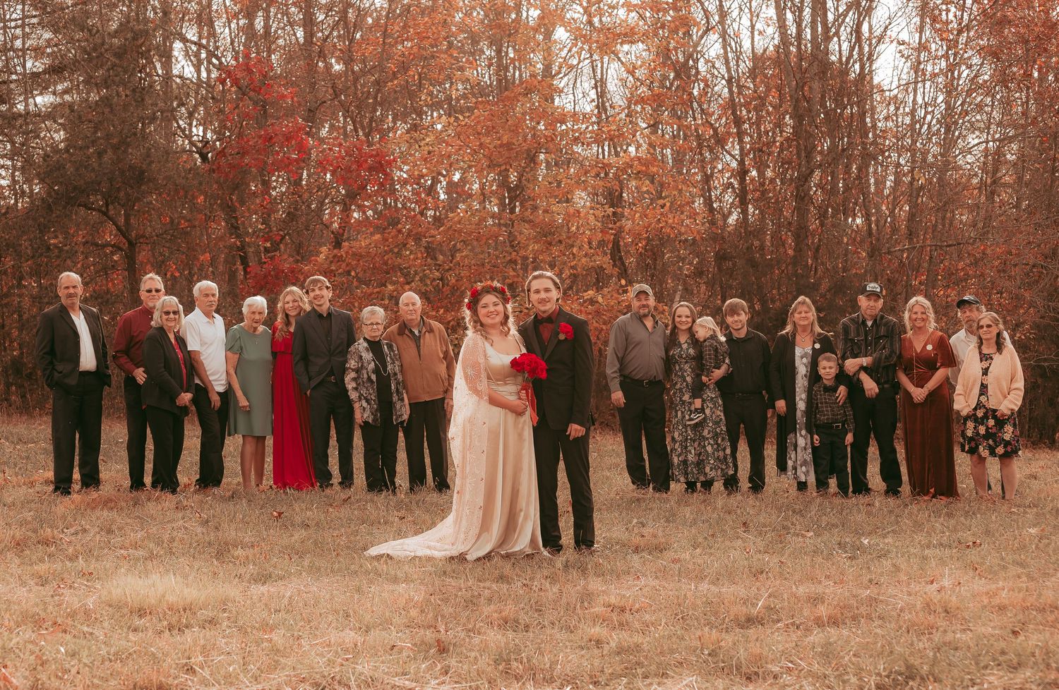 Fall wedding party portrait with bride in white gown and groom in black suit surrounded by family among autumn red foliage.