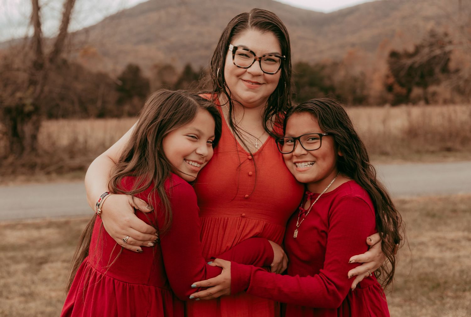 Mother in red dress hugging two daughters outdoors with mountain view background.