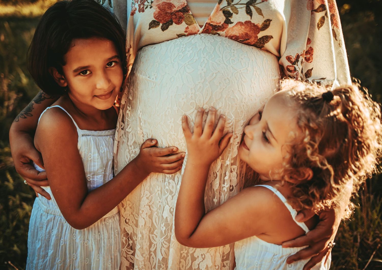 Two young girls hugging pregnant mothers lace-covered baby bump in golden afternoon light.