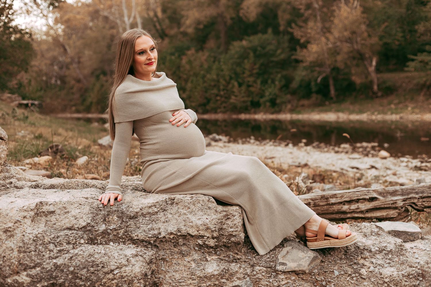 Elegant pregnant woman in beige maxi dress sitting on rocks near a peaceful river.