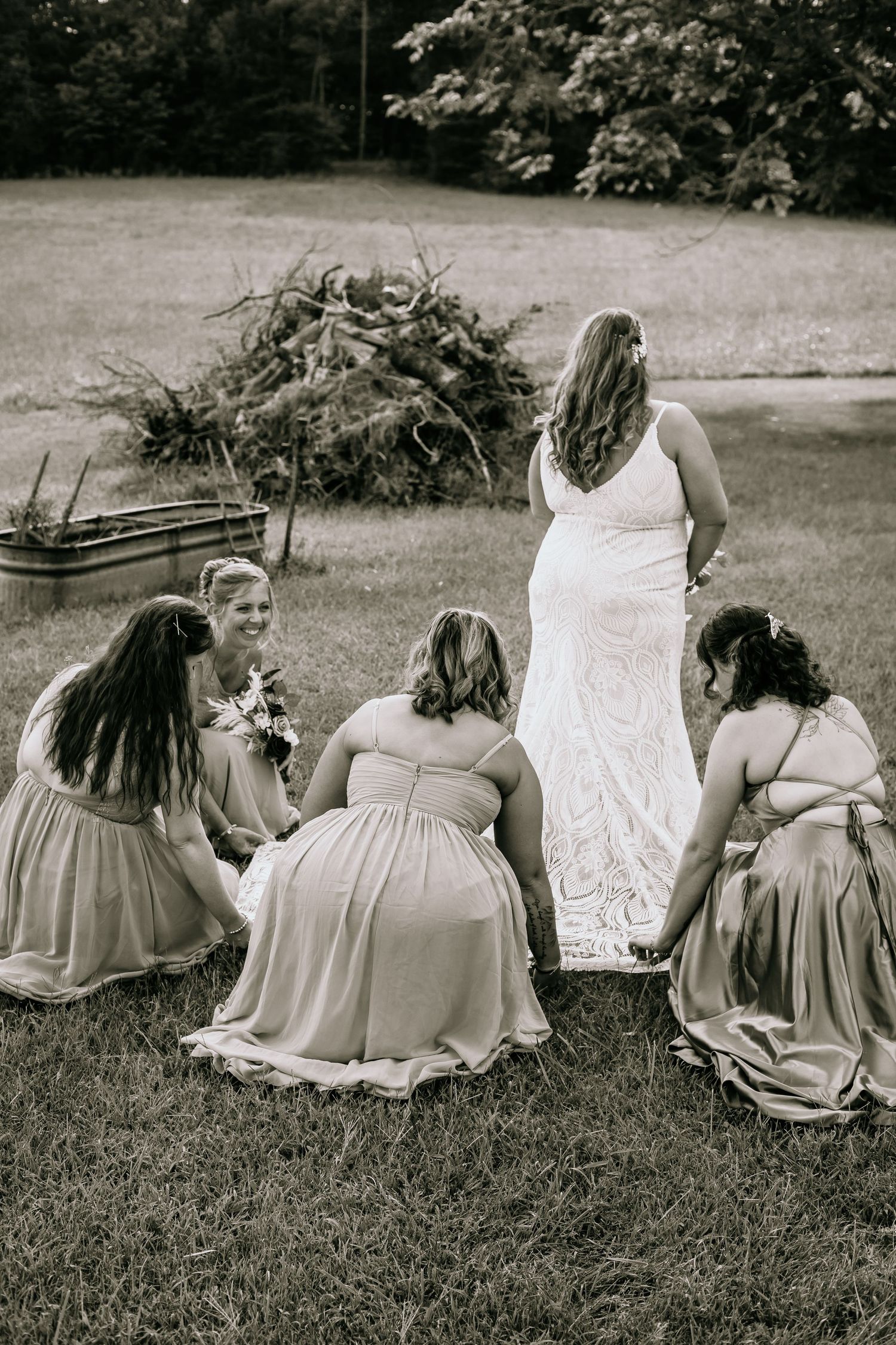 Black and white photo of bridesmaids adjusting the bride's lace wedding gown train outdoors.
