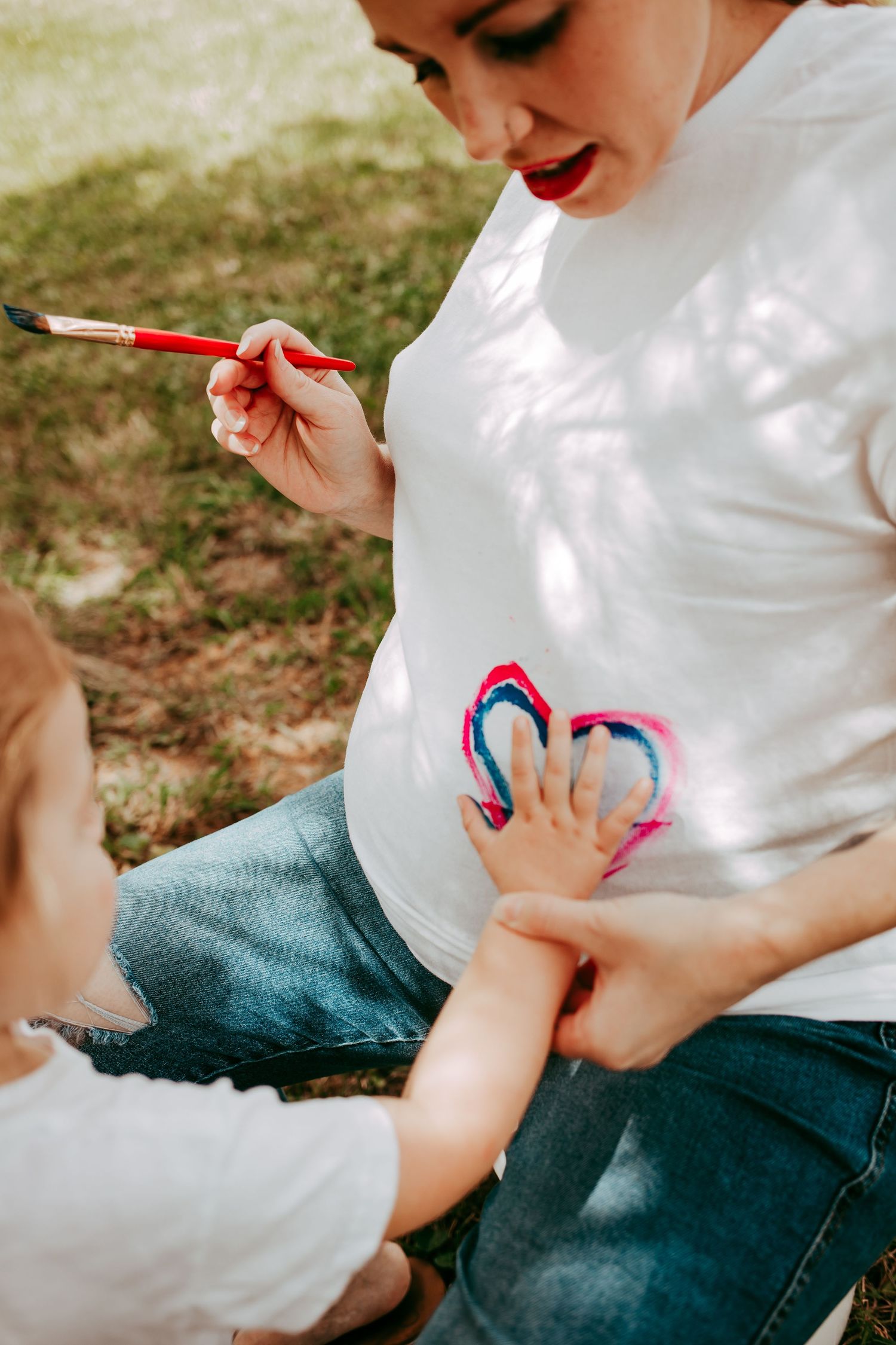 A pregnant mom and toddler paint a colorful heart design on her white maternity shirt in a sunny park.