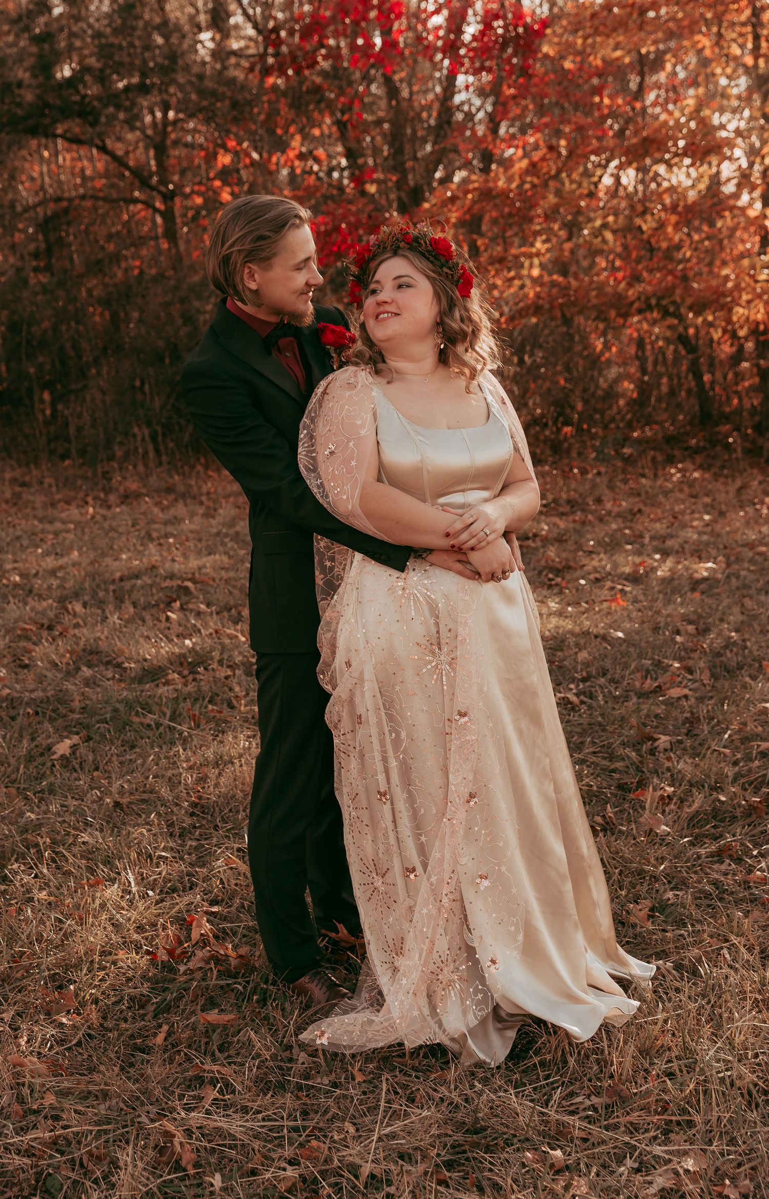 Groom embraces bride in gold lace wedding gown among vibrant autumn red foliage.