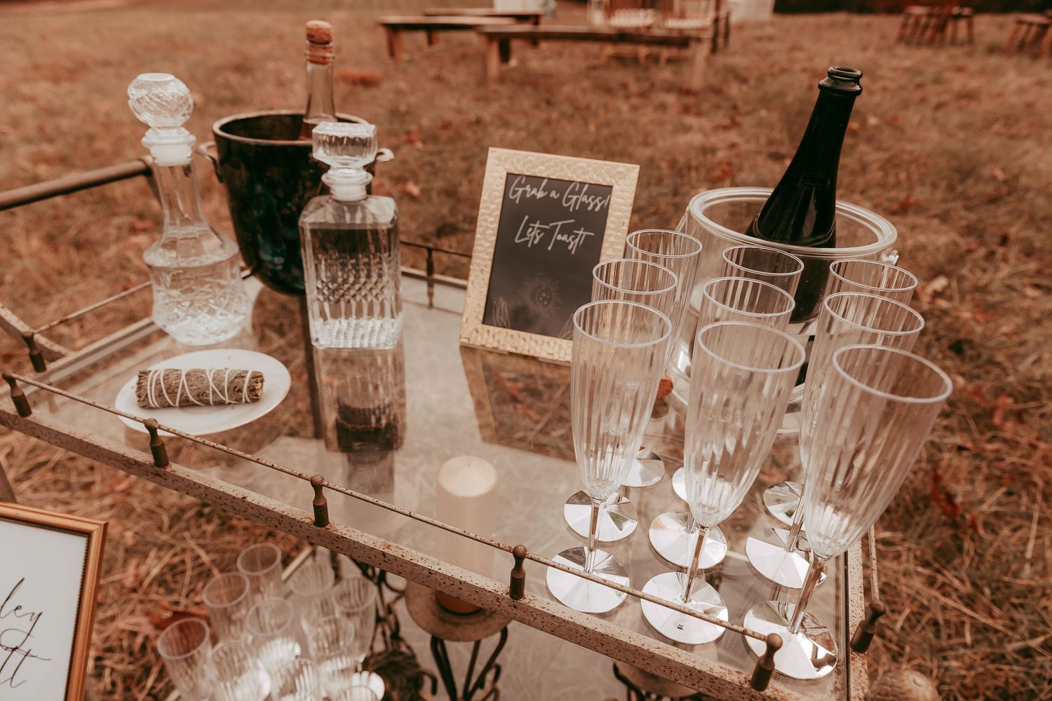 Elegant outdoor wedding champagne bar with crystal flutes, decanters, and chilled bottles on a mirrored tray in a fall field.