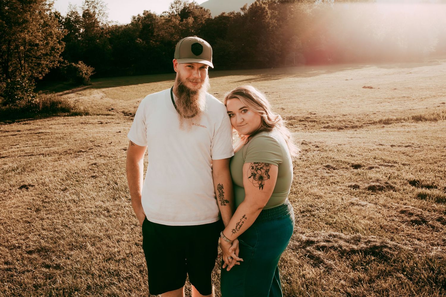 Tattooed couple holding hands and posing together in warm sunset light in open field with trees behind.
