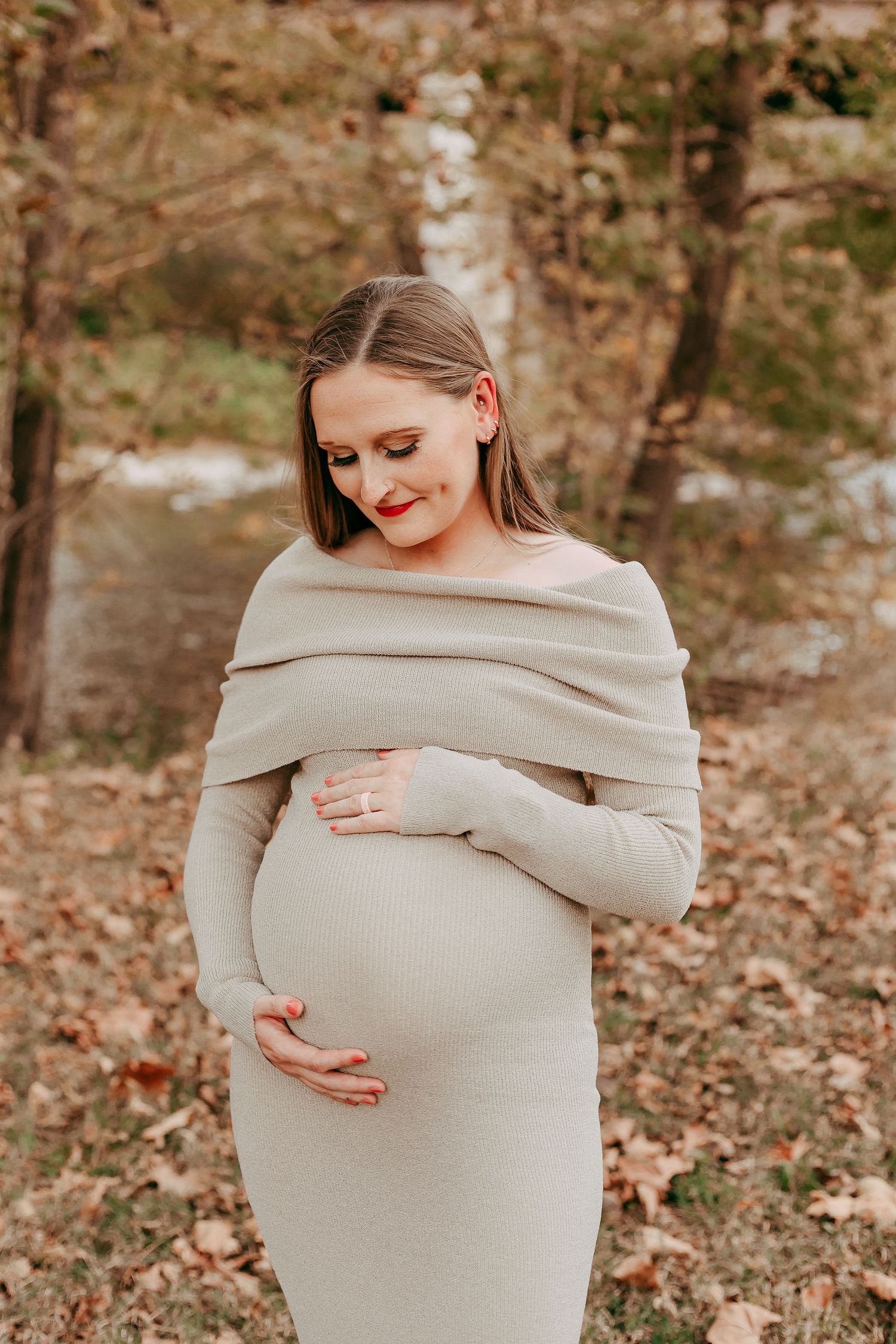 Pregnant woman in beige knit dress cradling baby bump among autumn leaves outdoors.