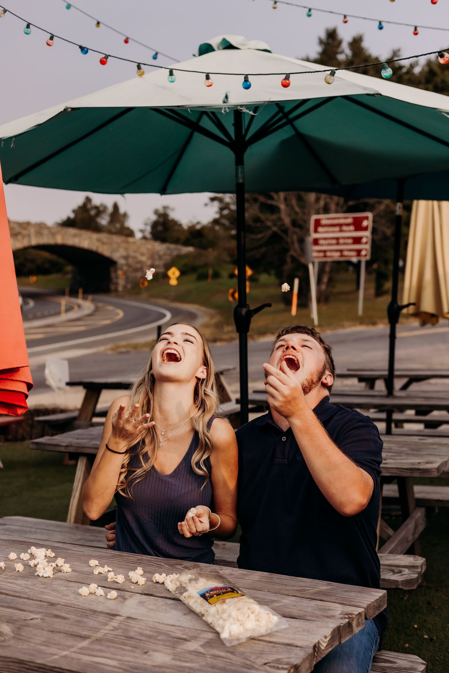 Couple laughing and catching popcorn in their mouths at an outdoor picnic table under string lights.