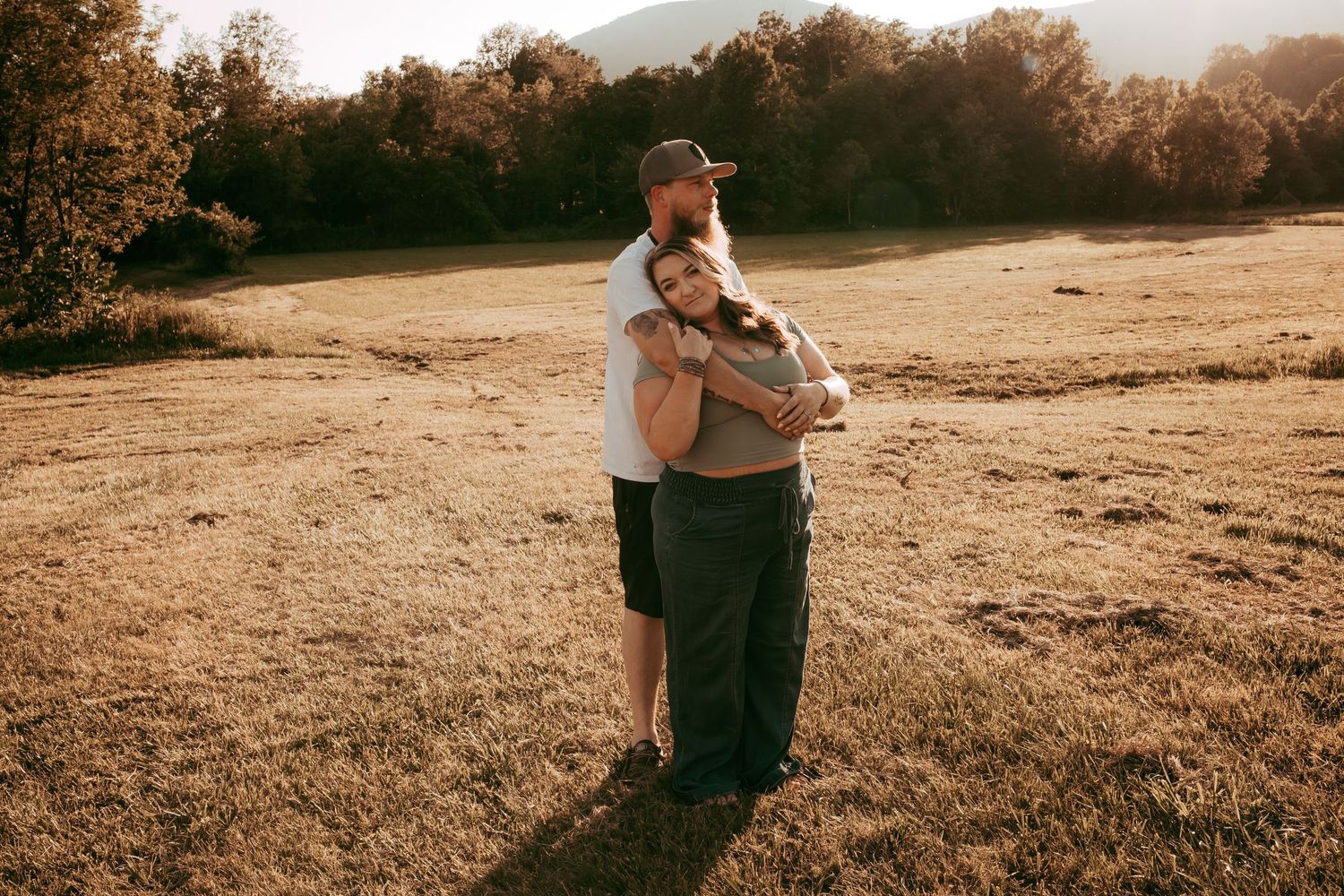 Couple sharing tender embrace in golden sunlit meadow with rolling hills and autumn trees in background.