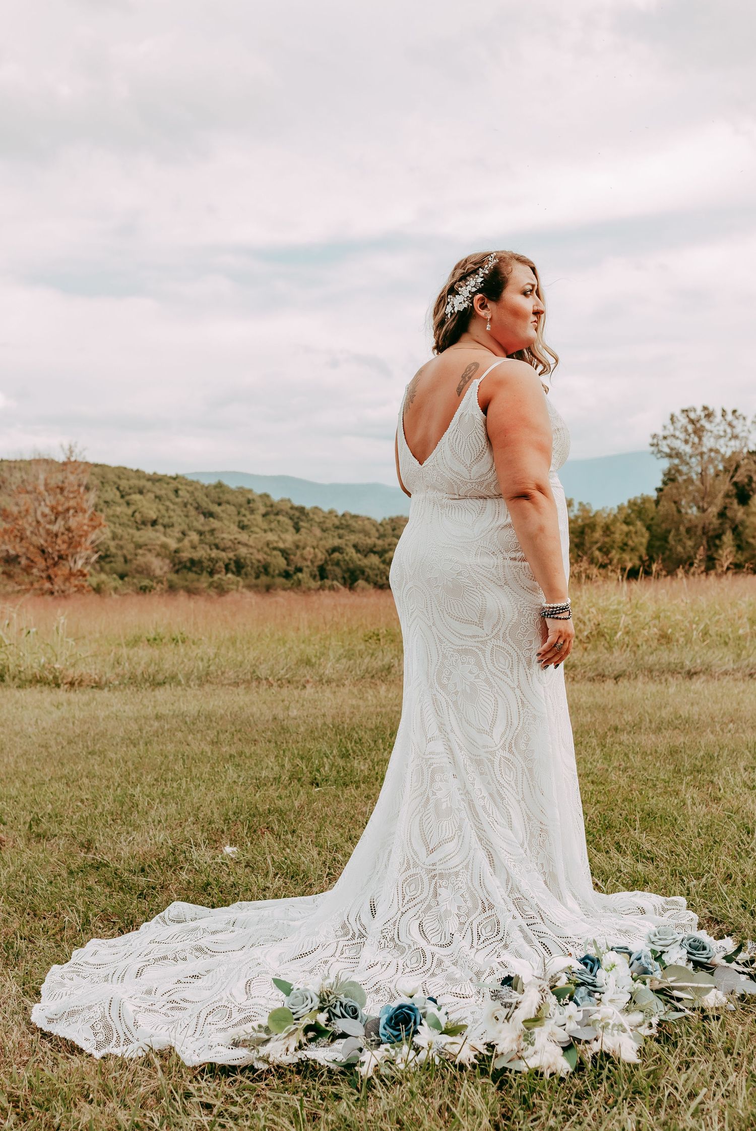 Bride in elegant lace gown with long train poses in a scenic mountain meadow on her wedding day.