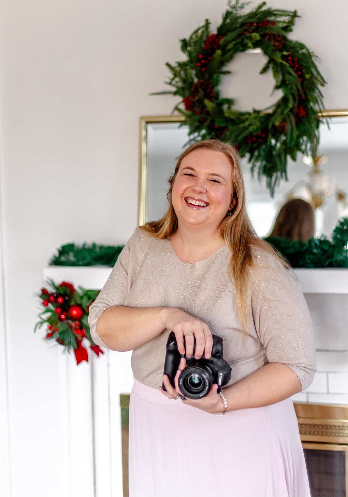 Smiling female photographer holding a camera in front of a Christmas fireplace with wreath and holiday decorations.