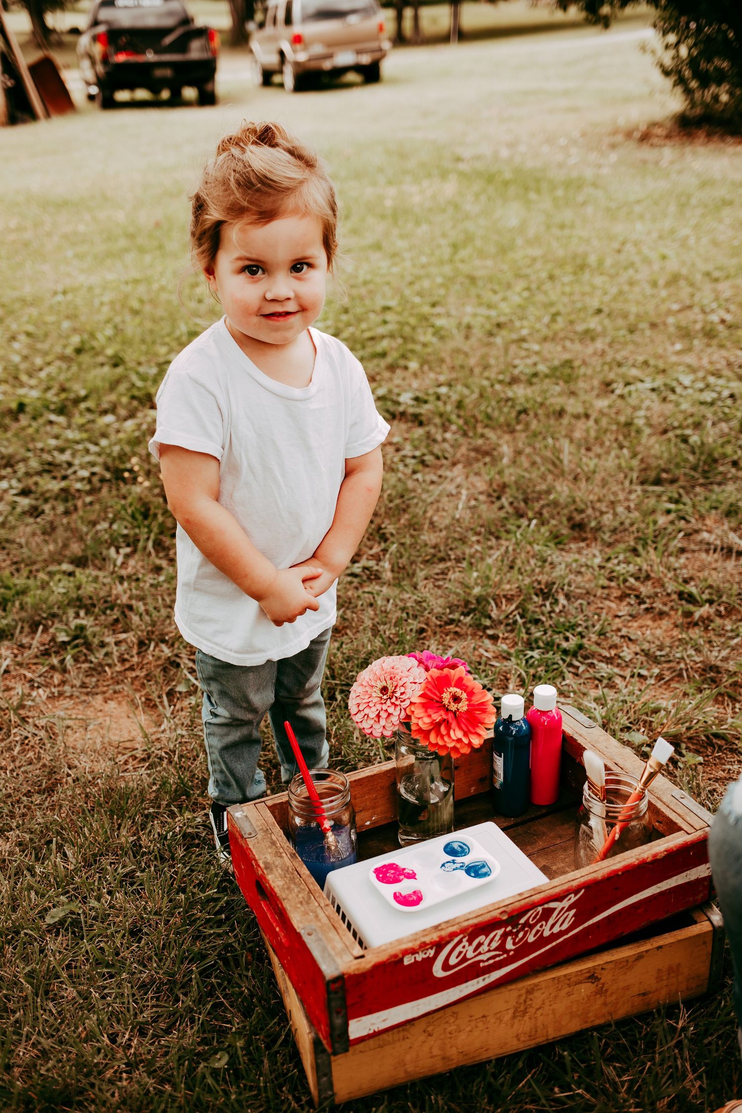 A young toddler in a white shirt stands beside a vintage Coca-Cola crate filled with paints and flowers outdoors.