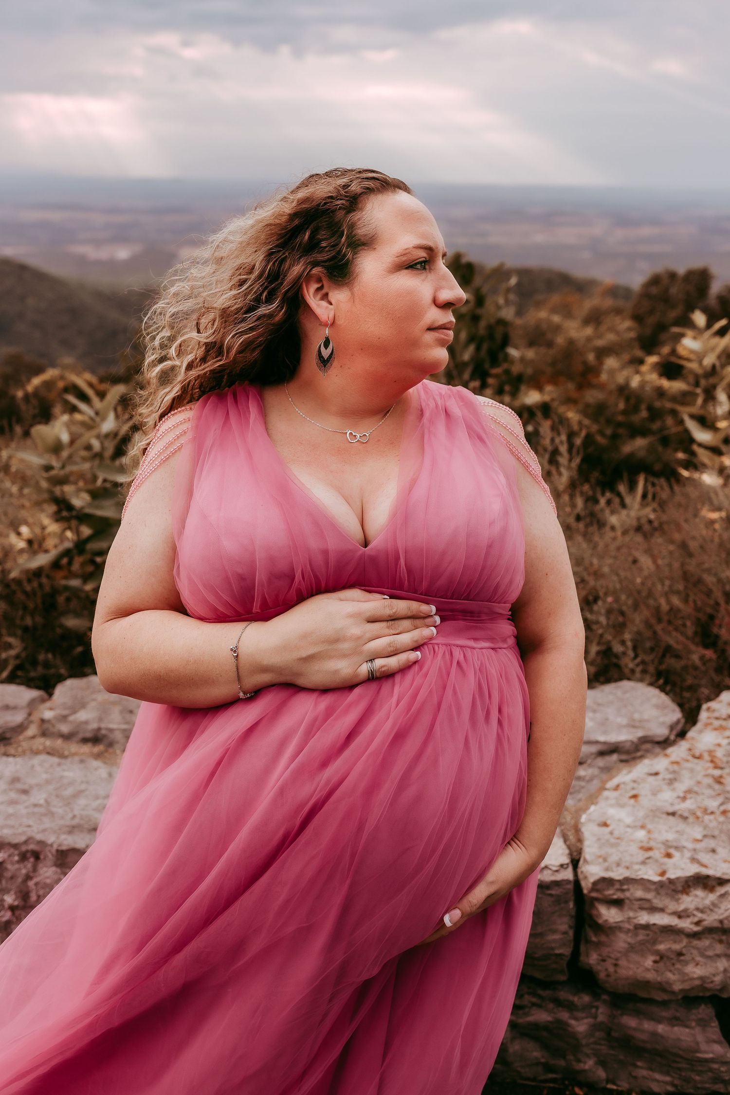 Pregnant woman in pink tulle dress at mountain overlook, holding baby bump with curly hair.