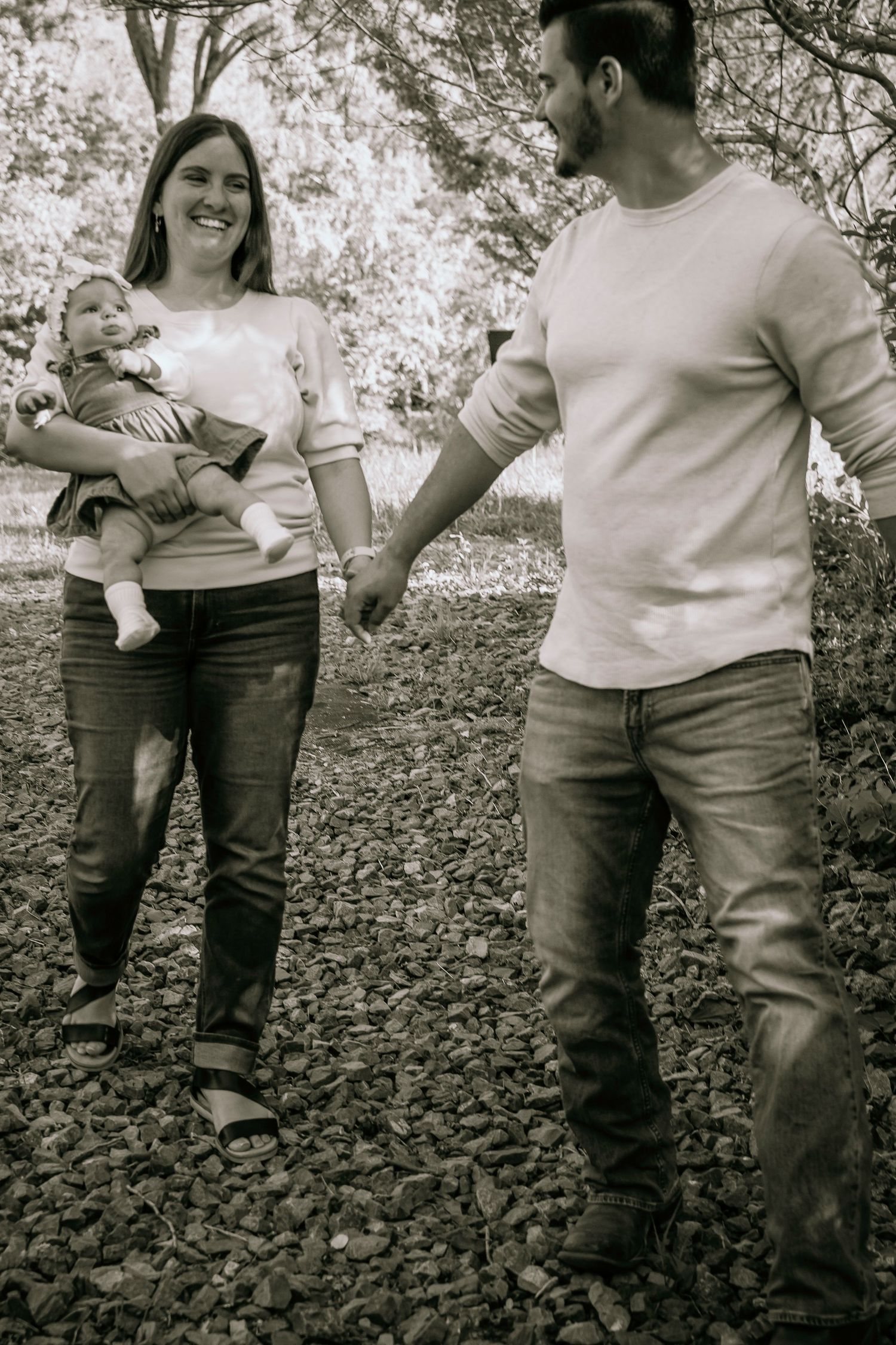 Black and white photo of a smiling couple holding hands outdoors, woman carrying a baby, standing on rocky ground near trees.