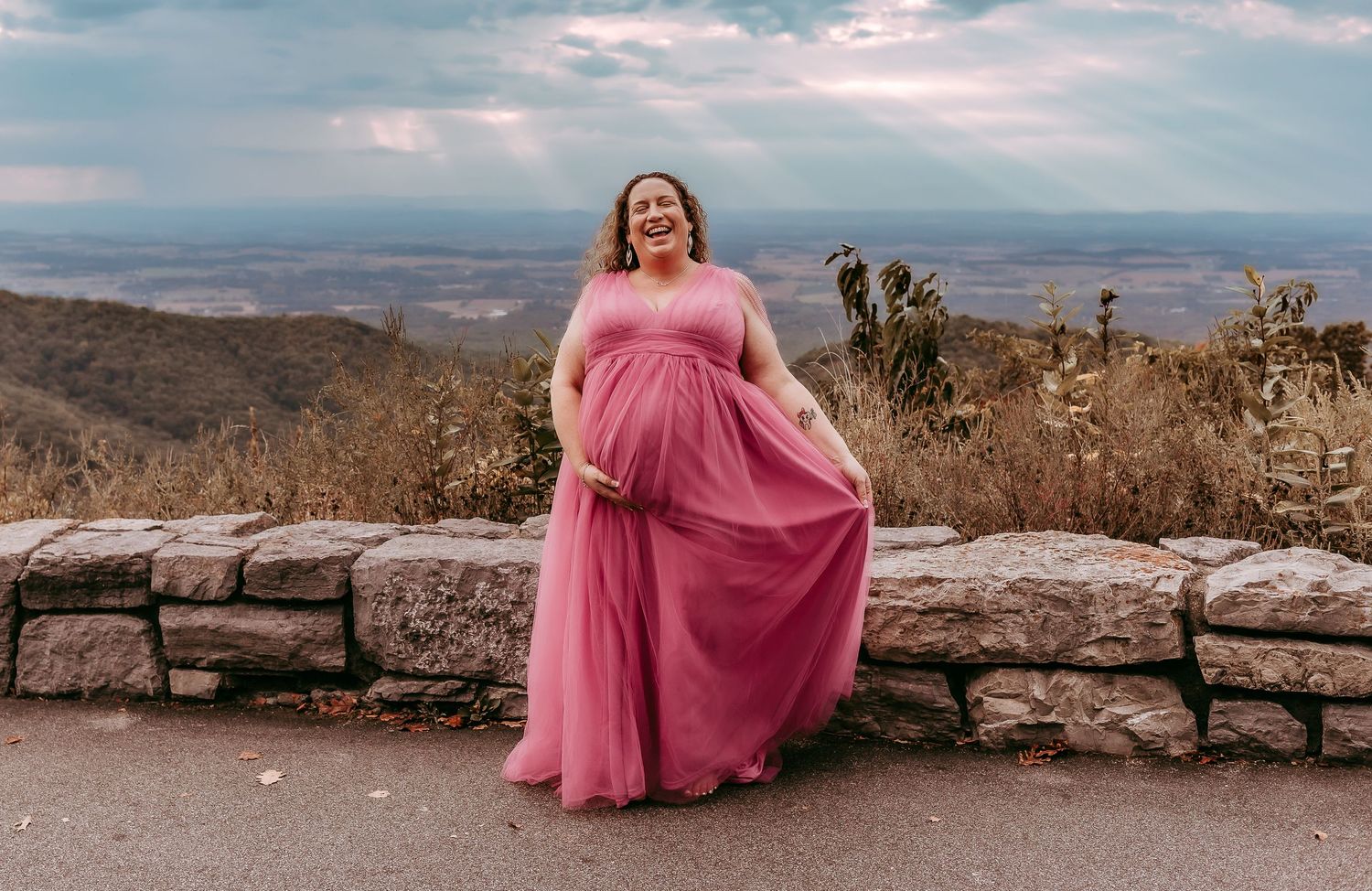 Pregnant woman in pink tulle gown laughing at mountain overlook with rays of sunlight.