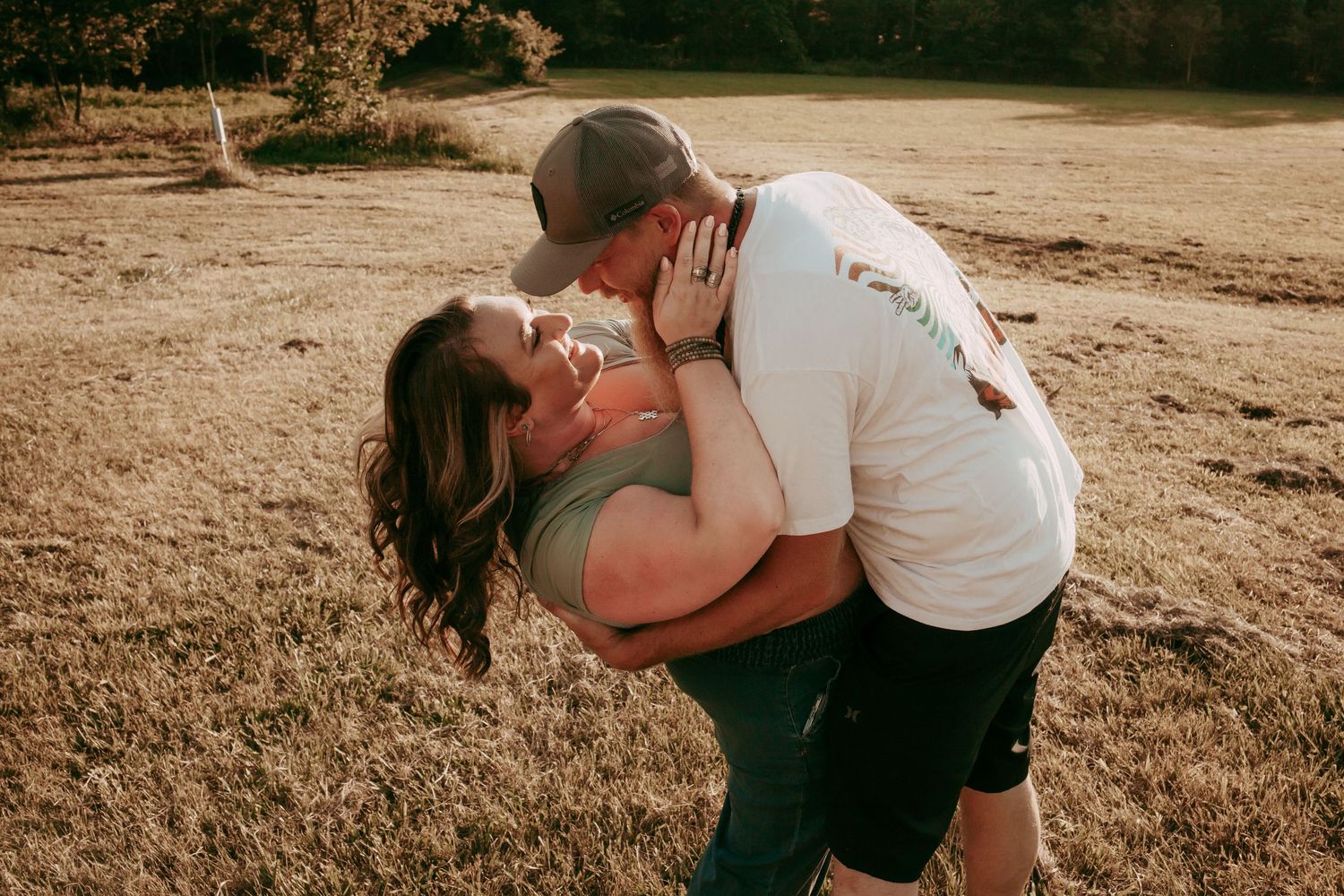 Couple sharing romantic dip kiss embrace in golden hour sunlit open field during outdoor photo session.