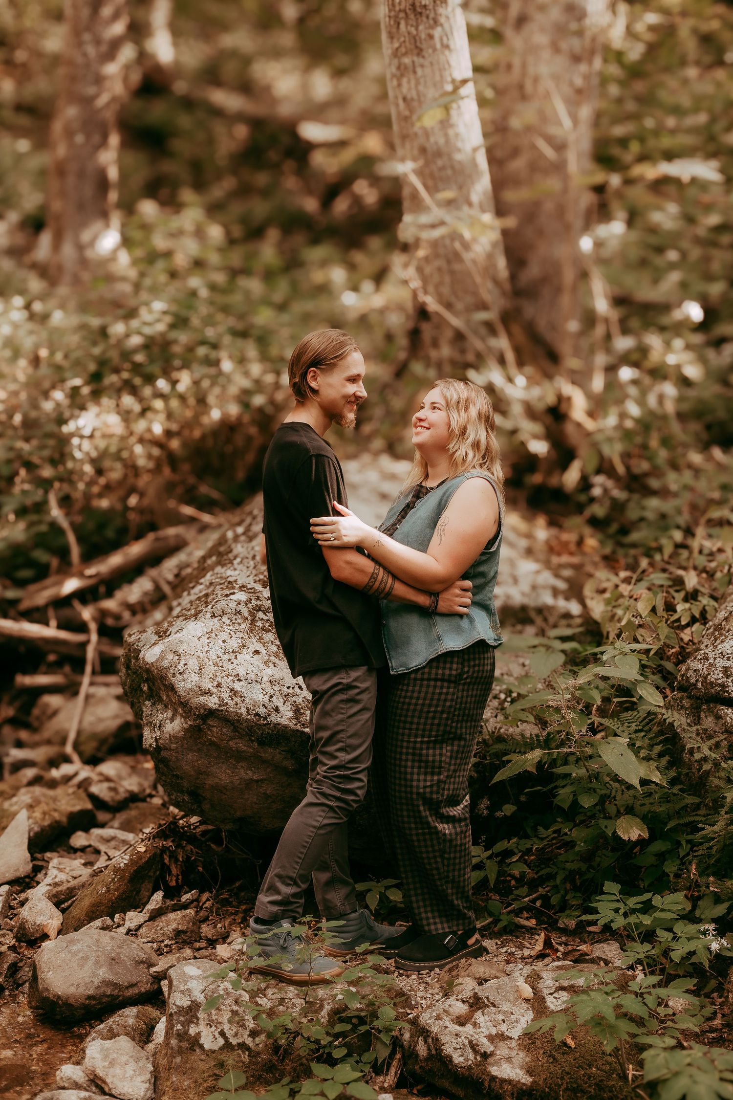Happy couple embracing and laughing together near large boulder in woodland setting.