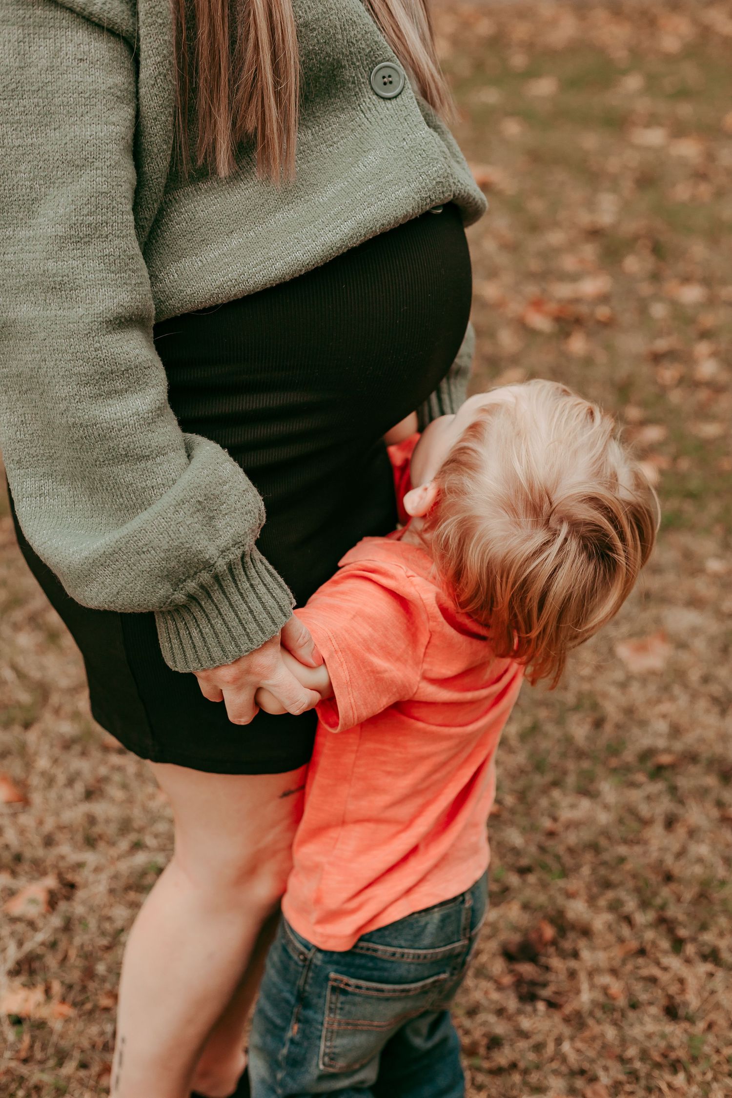 Close-up of toddler in orange shirt holding pregnant mothers hand outdoors in autumn.