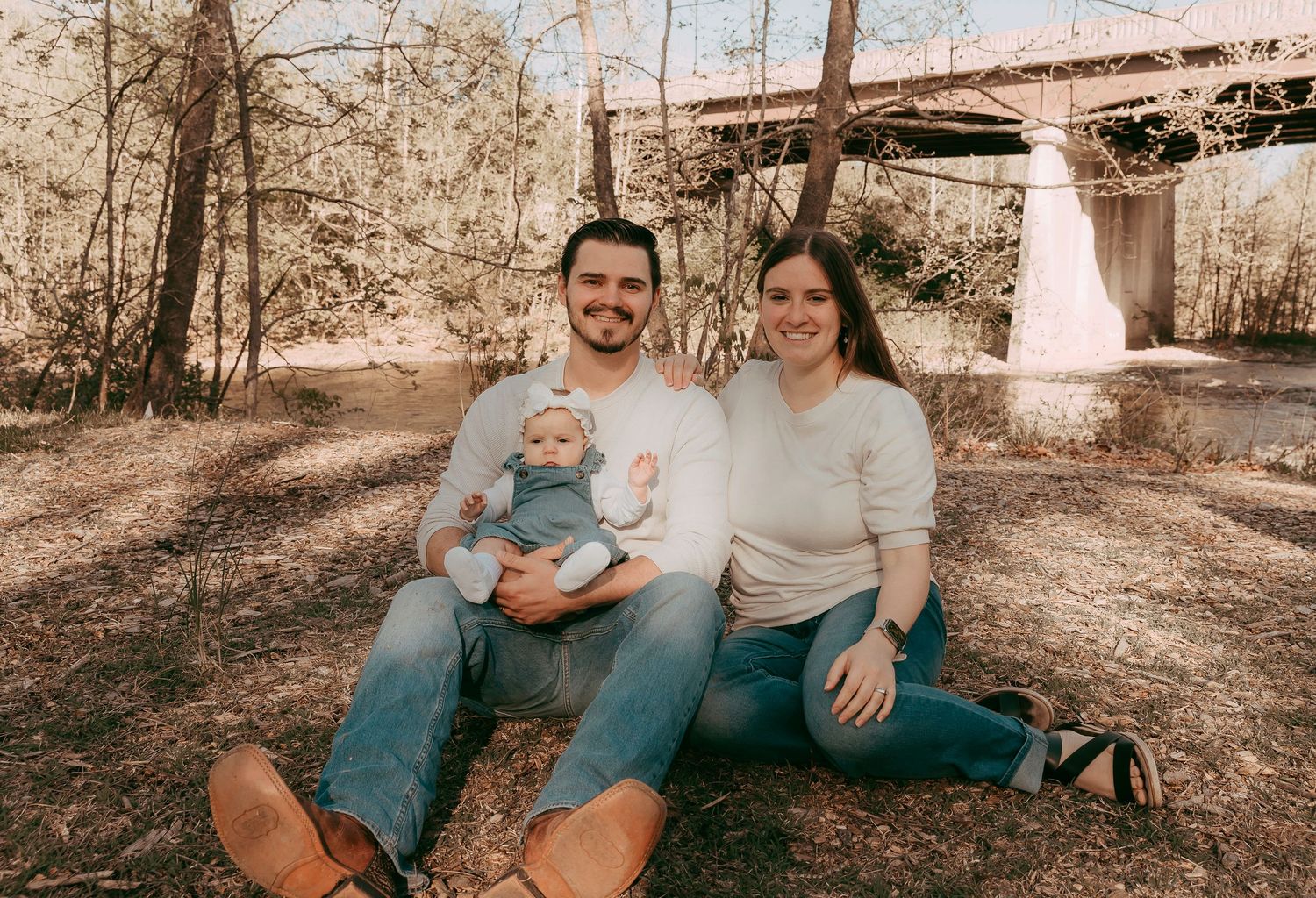 Young family with baby sitting outdoors near a bridge, smiling together in a wooded area.